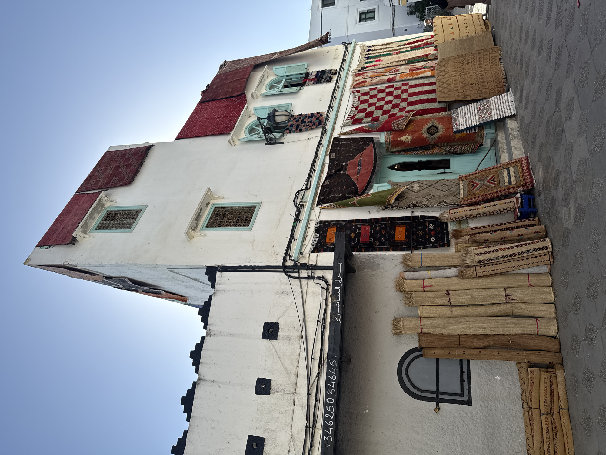 Captured from a low angle and rotated, the image portrays a sunlit building in Asilah, Morocco. The building's white facade serves as a display for a variety of rugs and carpets. The rugs come in different colors and patterns, including a red-and-white checkered rug, a brown rug with geometric patterns, and darker rugs with red and black details. Some rugs are hanging, while others are piled on the ground. The building also features arched windows with turquoise frames. A black sign on the wall displays white Arabic text, adding a local touch. The background includes a gray cobblestone street and a glimpse of other buildings. The sky is a clear blue. The overall impression is of a traditional Moroccan building that doubles as a carpet shop.