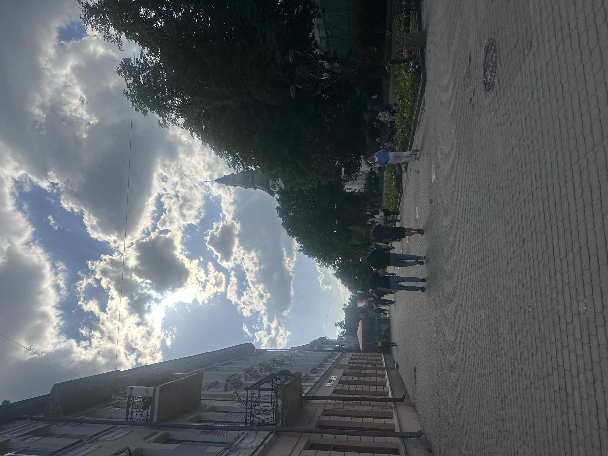 The image depicts a sunny day on a cobblestone street in Ternopil, Ukraine. In the foreground, a paved walkway is lined with buildings on the left and trees and a park area on the right. Several people are walking along the street, some individually and others in small groups. The architecture of the buildings suggests an urban setting, with multiple stories and balconies visible. Tall trees cast shadows on the walkway and park area. In the distance, a church steeple with a cross is visible, peeking above the trees. The sky is partly cloudy, with bright sunlight filtering through the clouds. The overall scene is one of a pleasant day in a city with historical architecture and green spaces.