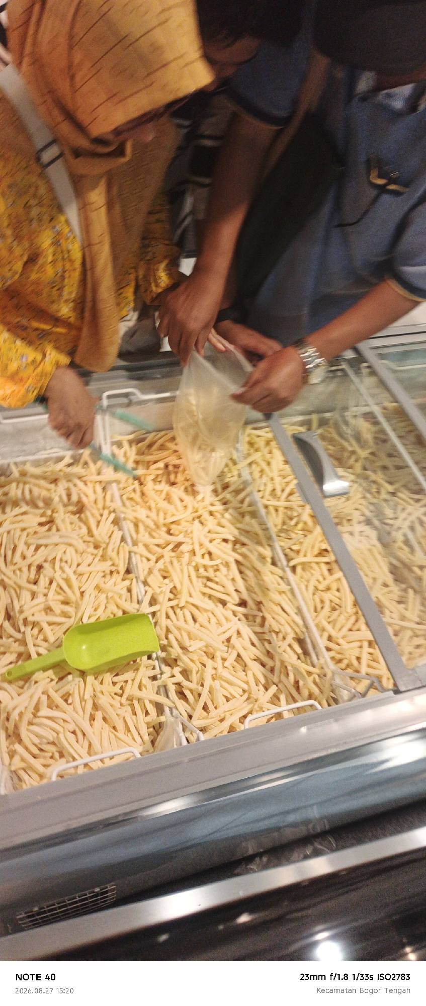 In a commercial freezer display case filled with french fries, two individuals are seen interacting. One person, wearing a yellow patterned garment, is partially visible on the left, reaching into the display. The other individual, wearing a blue shirt and a watch on their left wrist, is on the right, holding a clear plastic bag filled with french fries. They appear to be in the process of scooping or transferring the fries. A small, bright green plastic scoop is also visible in the display case, resting on the french fries. The scene suggests a food service or retail environment, possibly a restaurant or a market. The image is watermarked with "NOTE 40 2026.08.27 15:20" and "23mm f/1.8 1/33s ISO2783 Kecamatan Bogor Tengah," indicating the location as Bogor, Indonesia, and providing technical camera details.