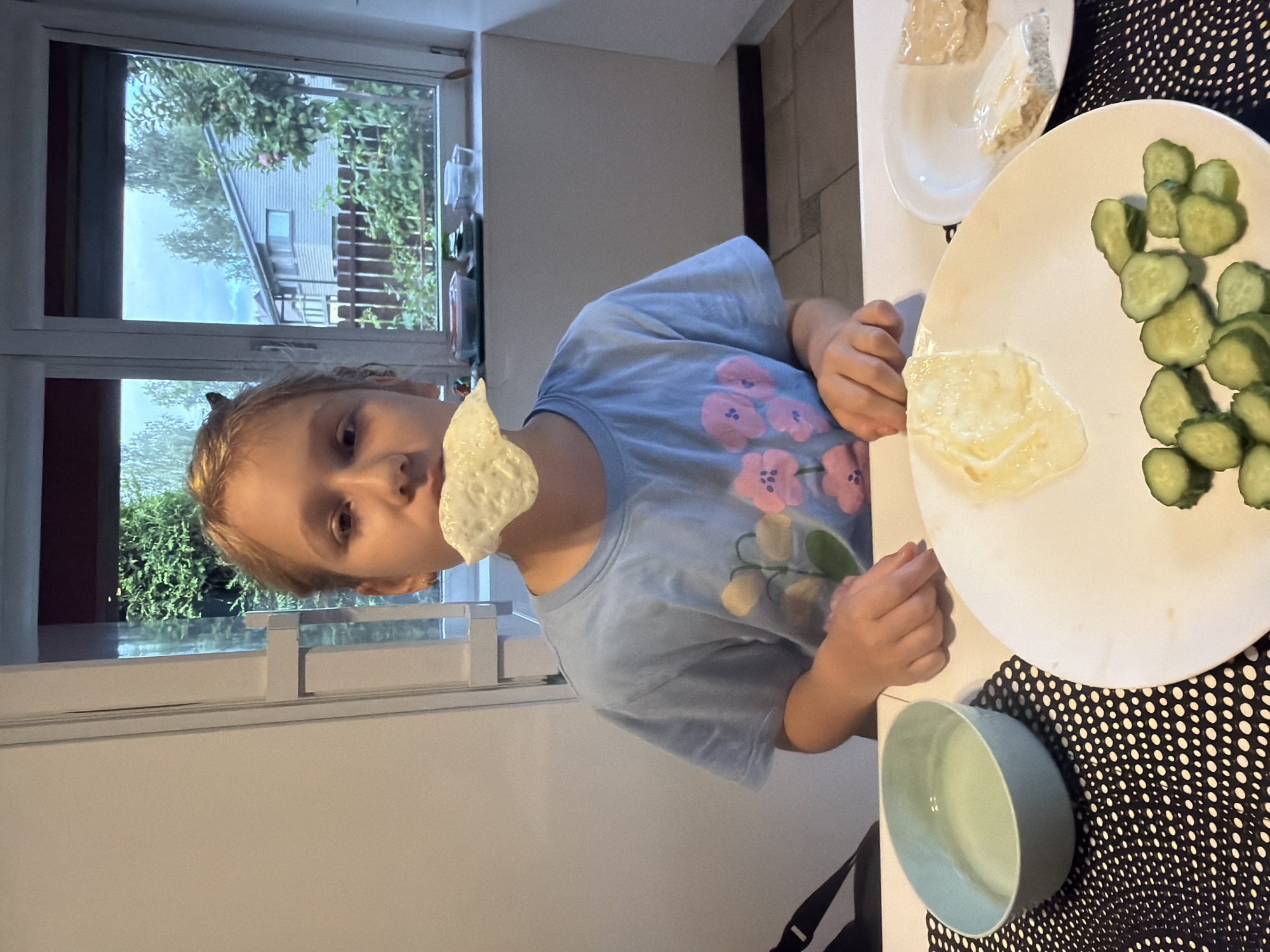 In an indoor setting, a young person with blond hair and a light blue t-shirt adorned with a floral pattern is seated at a table. They are holding a piece of food in their mouth. In front of them, a white plate contains sliced cucumbers and a portion of what appears to be scrambled eggs or a similar dish. To the right, another white plate holds two pieces of bread with a spread. To the left, a light green bowl contains water. A window in the background reveals a view of houses and trees, suggesting a residential area, potentially in Baloži, Latvia. The lighting indicates it is daytime.