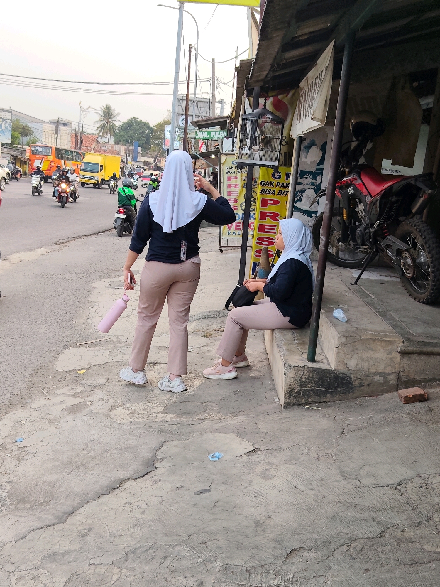 The image depicts a street scene in Purwakarta, Indonesia, characterized by moderate traffic and pedestrian activity. Two women, both wearing hijabs and casual attire, are the primary subjects. One woman, standing on the left, wears a dark long-sleeved shirt and light brown pants, and carries a pink water bottle. The other woman is seated on a concrete step to the right, wearing a similar outfit and has a black bag beside her.
The environment is a roadside with a paved street showing signs of wear and tear. Various vehicles, including motorcycles, cars, and trucks, are visible on the street, indicating a functional transportation artery. Along the roadside, there are small businesses or stalls with signage. One stall has a red motorcycle parked in front of it. The overall atmosphere suggests a typical day in a busy urban or semi-urban area. The lighting indicates it is likely daytime, possibly late afternoon or early morning given the soft light.