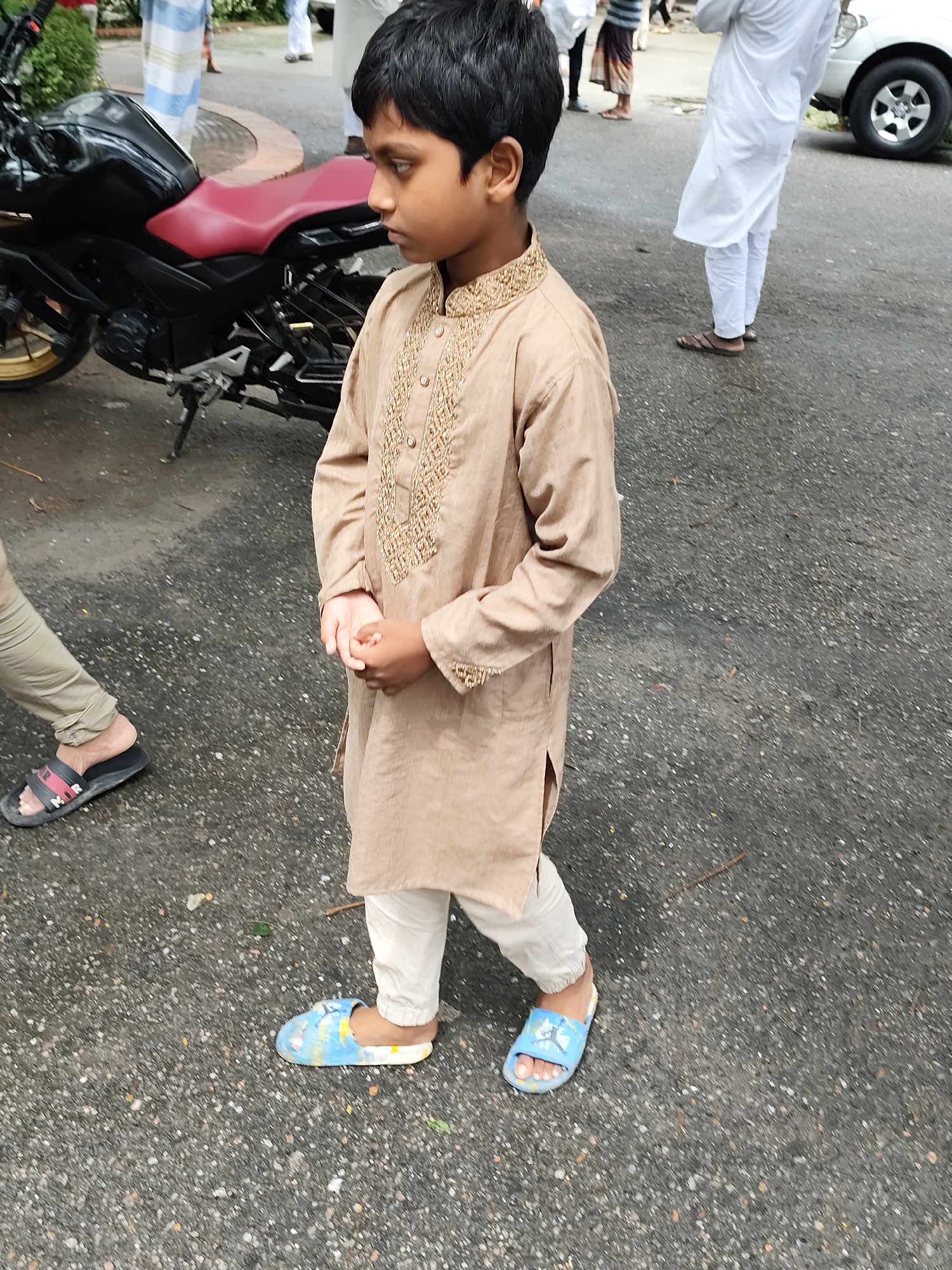 A young boy with dark hair and brown skin stands in profile on an asphalt surface. He is wearing a tan kurta with gold embroidery on the yoke and cuffs, and white pants. His feet are adorned with blue and white patterned slides. To the left of the boy, a black motorcycle is parked. In the background, two individuals are visible. One is wearing a white salwar kameez, and the other is wearing a white shirt and pants. The scene appears to be outdoors, with a hint of greenery to the left and a white SUV in the far background to the right. The overall impression is of a casual outdoor gathering in a city setting.