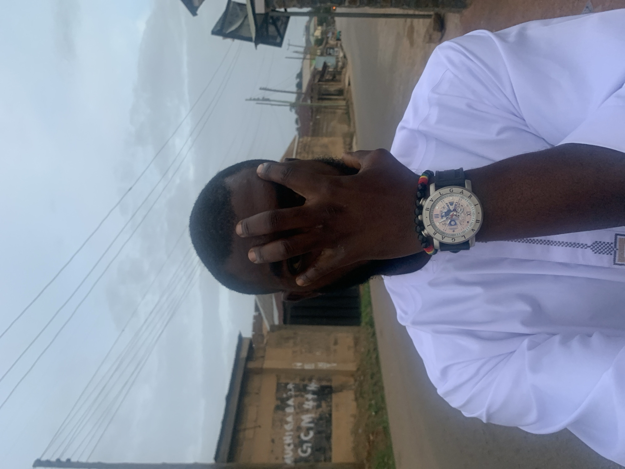 A man wearing a white shirt and a watch is standing on a street. He has his hand over his face. The background includes buildings and power lines, suggesting a city setting. On the wall of a building, some graffiti in white letters says "AUCHI ASA GCM 41+11". The location of the image is Jos, Nigeria.
