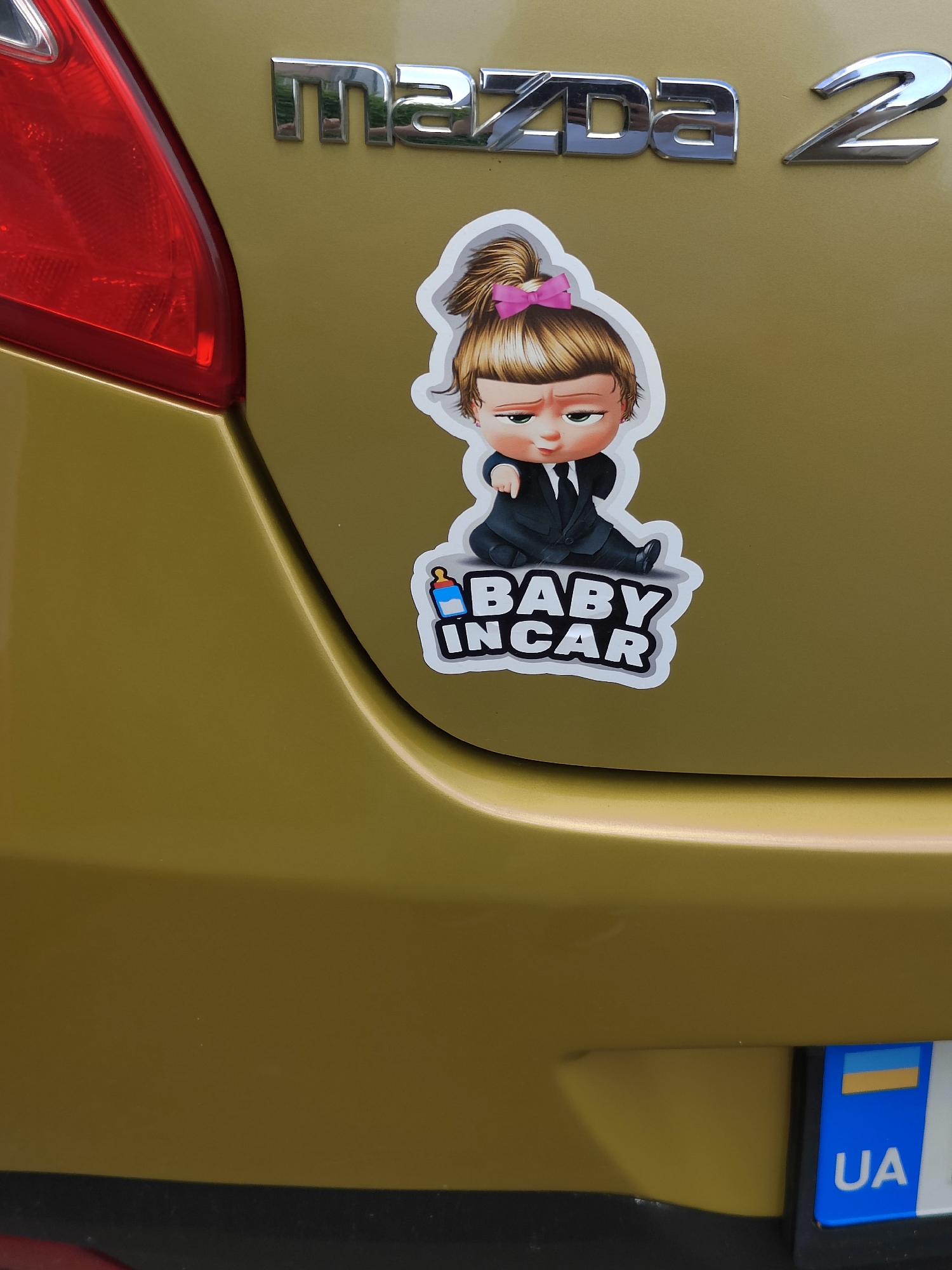 The image is a close-up shot of the rear of a gold Mazda 2 car parked outdoors. A sticker depicting a character from "The Boss Baby" franchise is prominently displayed on the car's trunk. The sticker shows the baby character wearing a suit and tie, with the words "BABY IN CAR" written below. The car also features a Ukrainian license plate. The lighting suggests it is daytime, with the sun illuminating the car's surface. No people are visible in the image. The car appears to be in a city environment, possibly on a street or in a parking area. The overall impression is of a well-maintained vehicle with a humorous personal touch.