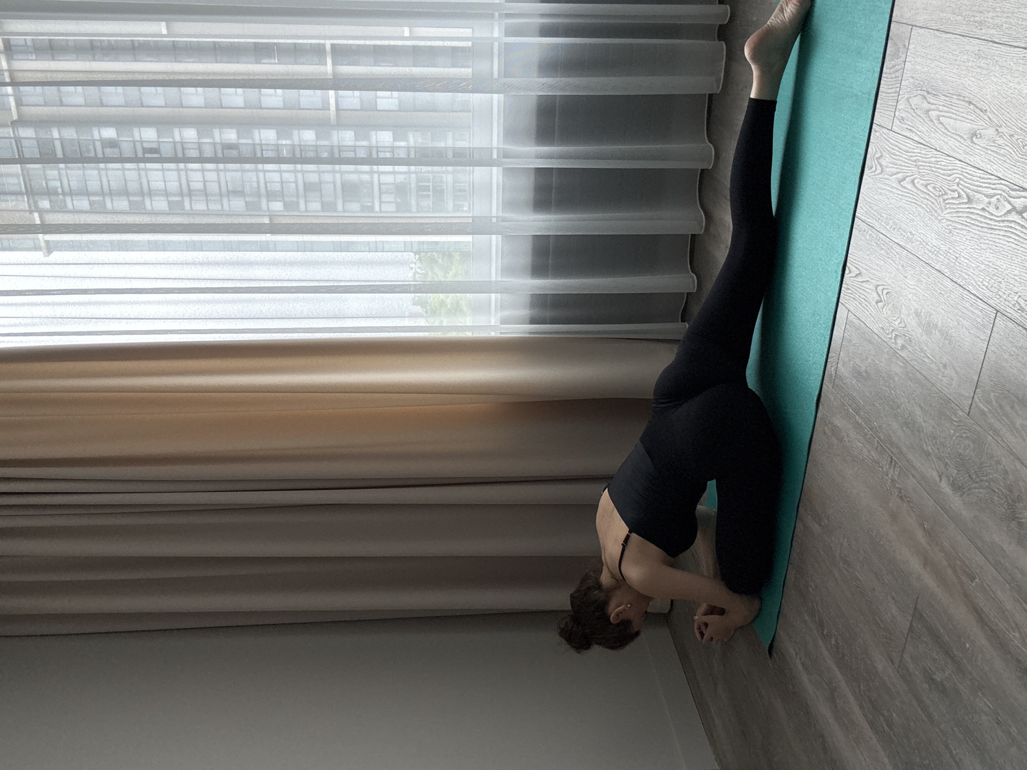 A woman is performing a yoga pose called Pigeon Pose on a turquoise yoga mat. She is wearing a black sports bra and black leggings. Her body is positioned with her right leg bent and positioned forward, while her left leg is extended straight back. Her upper body is lowered, with her forearms resting on the mat and her head bent down. She is in a room with light-colored wooden flooring and beige curtains on the left. A sheer white curtain is on the right, revealing a modern building visible through a window. The scene is likely indoors, in an apartment or studio, possibly in the city of Khimki, Russia, given the architecture visible outside the window.