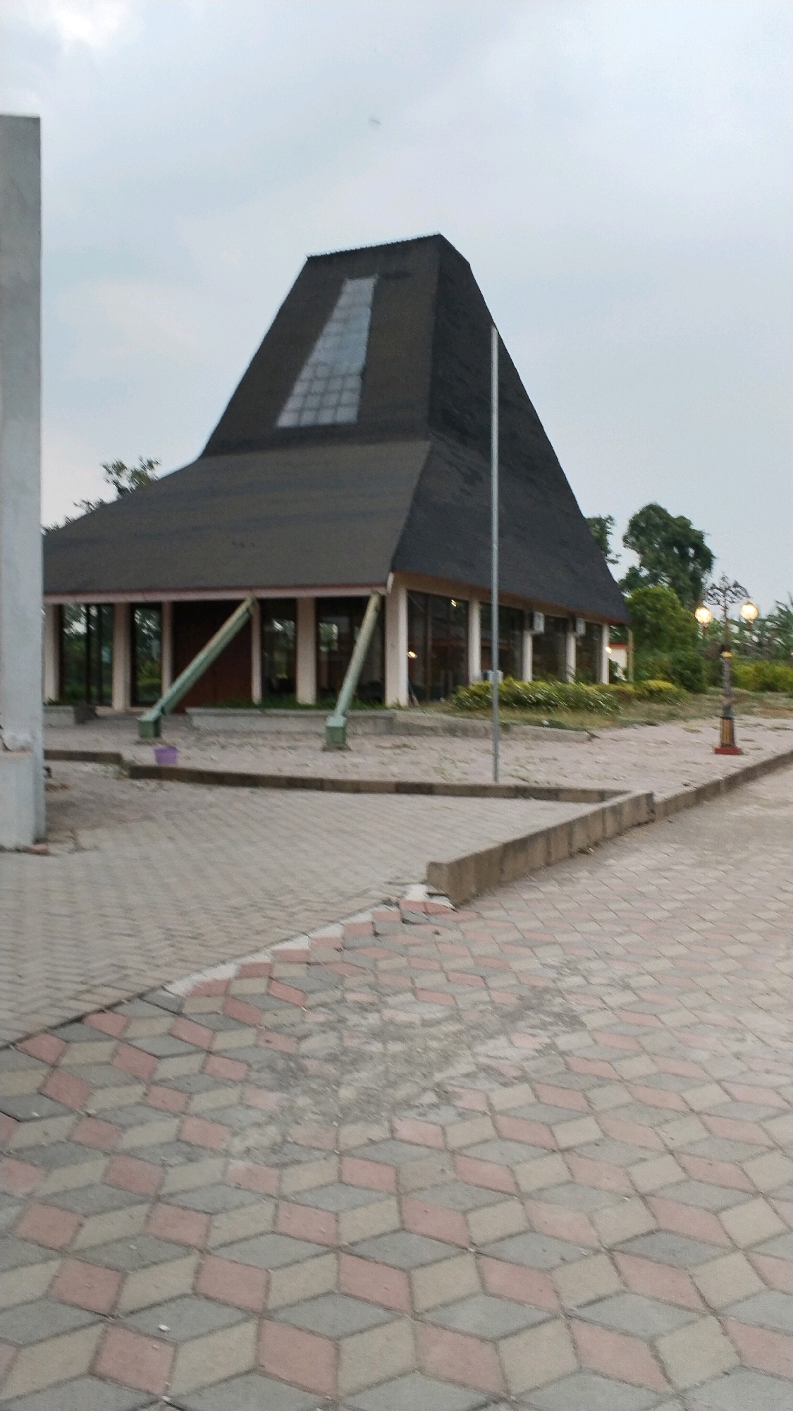 The image depicts a modern building with a dark, angular roof and a glass facade. The building is situated in an outdoor area with paved walkways. The paving stones in the foreground have a geometric, three-dimensional pattern. To the right of the building, a lamppost with two illuminated globes stands. The sky is overcast. The scene appears to be a public space, possibly a park or a community center, in Ngingasrembyong, Indonesia. There are no people visible in the image.