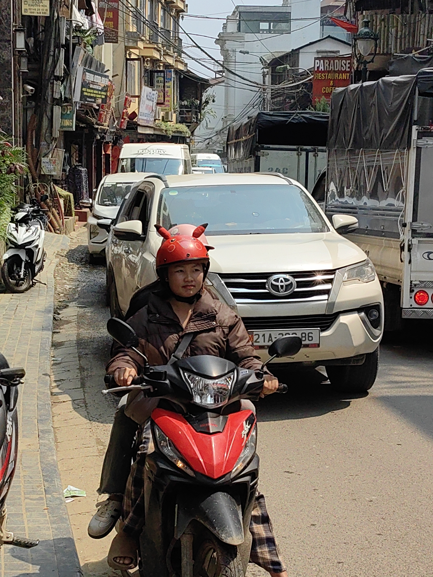 The image depicts a congested street scene in Sa Pa, Vietnam, viewed from a slightly elevated perspective. In the foreground, an individual wearing a brown puffer jacket, dark trousers, and a distinctive red helmet with prominent red "devil horn" protrusions, sits astride a red and black scooter, holding the handlebars. One foot, wearing an open-toed sandal, is visible resting on the ground or footrest.
Immediately in front of the scooter is a white Toyota Fortuner SUV, displaying a Vietnamese license plate partially visible as "21A-XXX.86". Behind the Fortuner, a line of vehicles includes another white SUV/van and a white passenger van with "LUXURY" branding. To the right of the Fortuner, a dark truck with a black tarp-covered bed is stationary, with the rear of another similar truck visible beside it.
To the left of the scooter, a raised sidewalk made of light-colored pavers runs alongside the street. A white Honda scooter is parked on this sidewalk, facing the viewer. Beyond it, various items, including what appear to be woven materials or stacked goods, are arranged against the facade of a multi-story building. This building, like others lining the street, features multiple signs, including "SAPA HOTEL" and "MASSAGE" in red, along with other Vietnamese text. Numerous electrical wires are visible crisscrossing overhead.
On the right side of the street, more multi-story buildings with balconies and windows are present. A prominent red sign advertises "PARADISE RESTAURANT & COFFEE." The street is paved with asphalt and appears to be in an urban setting, possibly on a slight incline. The scene is illuminated by daylight, indicating clear weather.