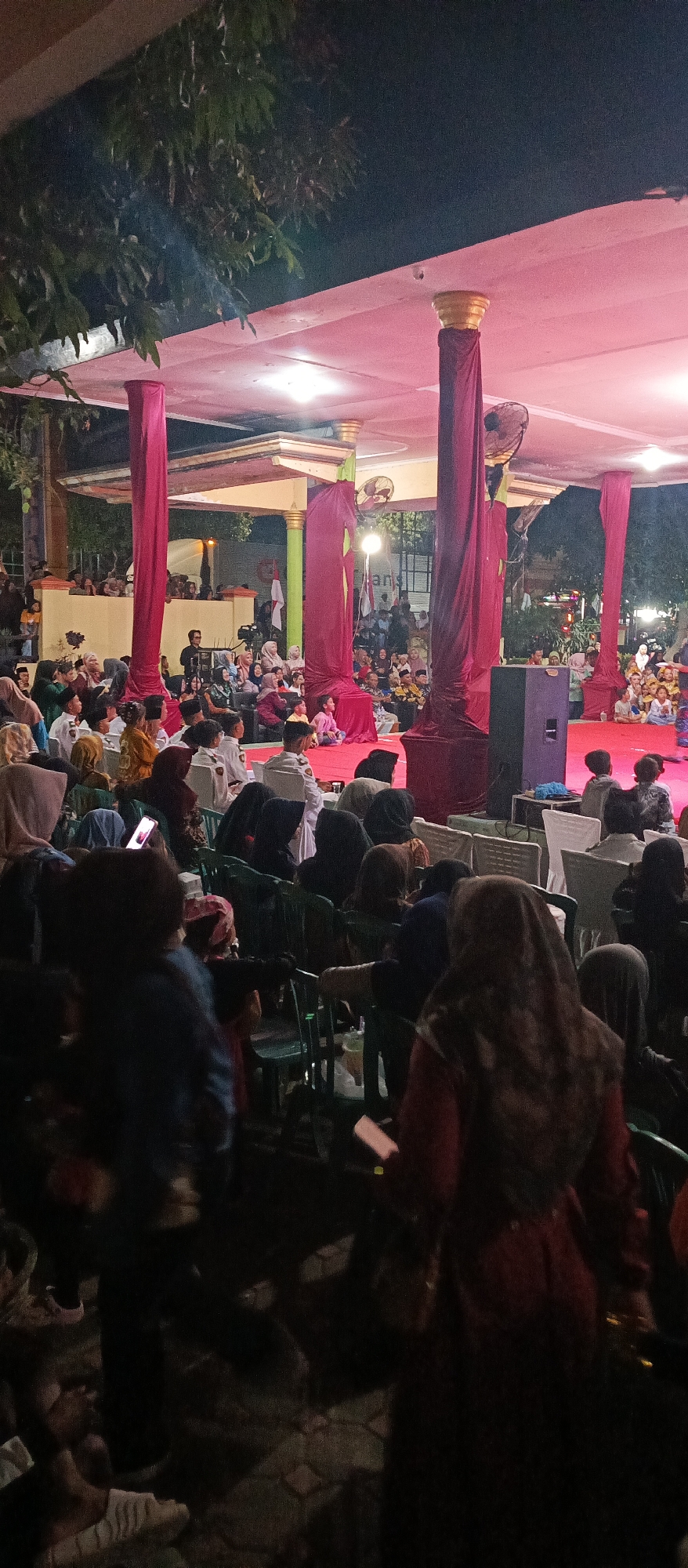 The image depicts an outdoor gathering at night, possibly an event or performance, in Arjasa, Indonesia. A stage area is visible, draped with deep red fabric and illuminated by pink lighting, with large golden-topped columns supporting a covered structure. In the foreground and midground, numerous people are seated on chairs, facing the stage, indicating they are an audience. Many individuals are wearing headscarves, common attire in Indonesia. The audience appears to be composed of adults and children. Some are looking towards the stage, while others are engaged in conversation or looking at their phones. In the background, more people are visible, some standing and others seated. The environment suggests a public space, possibly a community center or an open area designated for events. The overall atmosphere seems to be one of communal engagement, with the red drapes and lighting adding a festive or ceremonial touch to the setting.