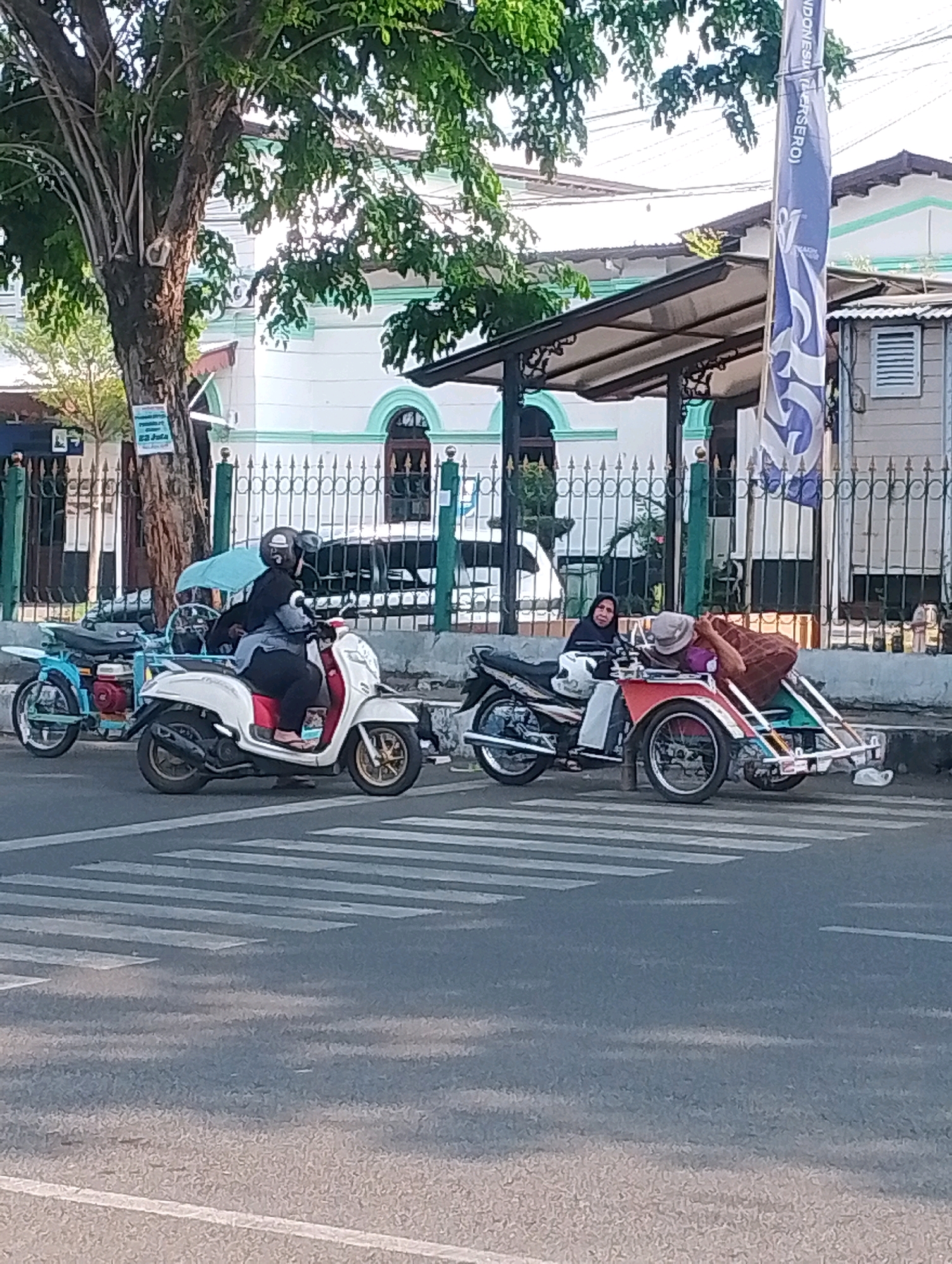 In the city of Probolinggo, Indonesia, a street scene unfolds near a building with white walls and green trim. A pedestrian crossing marked by white stripes dominates the foreground. On the street, a white scooter with red accents is parked, ridden by a person wearing a black helmet and dark clothing. Behind it, a blue motorcycle with a red engine is also parked. To the right, a motorcycle with a sidecar is positioned, with one person on the motorcycle and another reclining in the sidecar, covered by a brown and red patterned blanket and wearing a hat. The background shows a wrought-iron fence, a sidewalk, and a portion of a building. A tall tree with lush green leaves stands to the left of the frame.