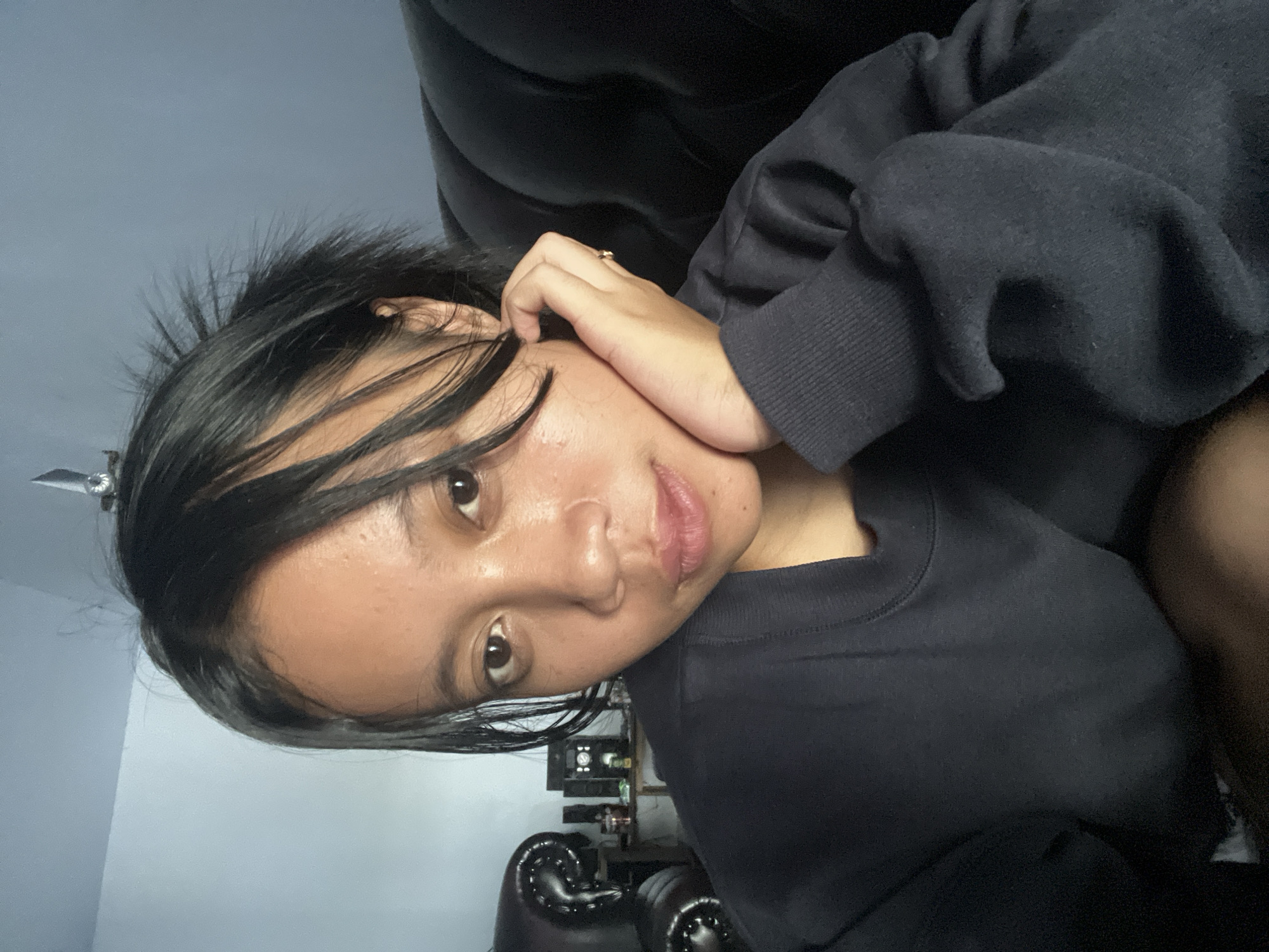 A young female with dark hair is captured in a close-up, selfie-style portrait. She is leaning her right cheek on her right hand, which is adorned with a simple ring on her ring finger. Her hair is somewhat disheveled, with strands falling across her face and forehead. She is wearing a dark, crew-neck sweatshirt. The background features a dark, upholstered couch on the right and a light blue wall on the left, with some shelves and objects visible on the wall. The scene appears to be indoors, possibly a living room. The lighting is dim, suggesting it might be evening or a low-light indoor environment. The woman's expression is neutral to slightly pensive. The image quality is moderate, with some visible texture and grain.