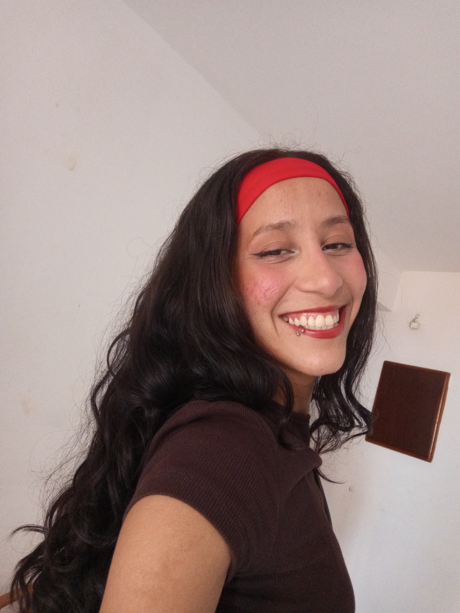 A smiling person with long, dark, wavy hair and a red headband is looking at the camera. They are wearing a brown top. The person has a lip piercing, visible red lipstick, and heavy blush. The background is a white wall with a dark brown, rectangular object attached to it, possibly a tile or panel. The location is not discernible based on the image content. The image does not contain any information indicating it was taken in Porlamar, Venezuela.