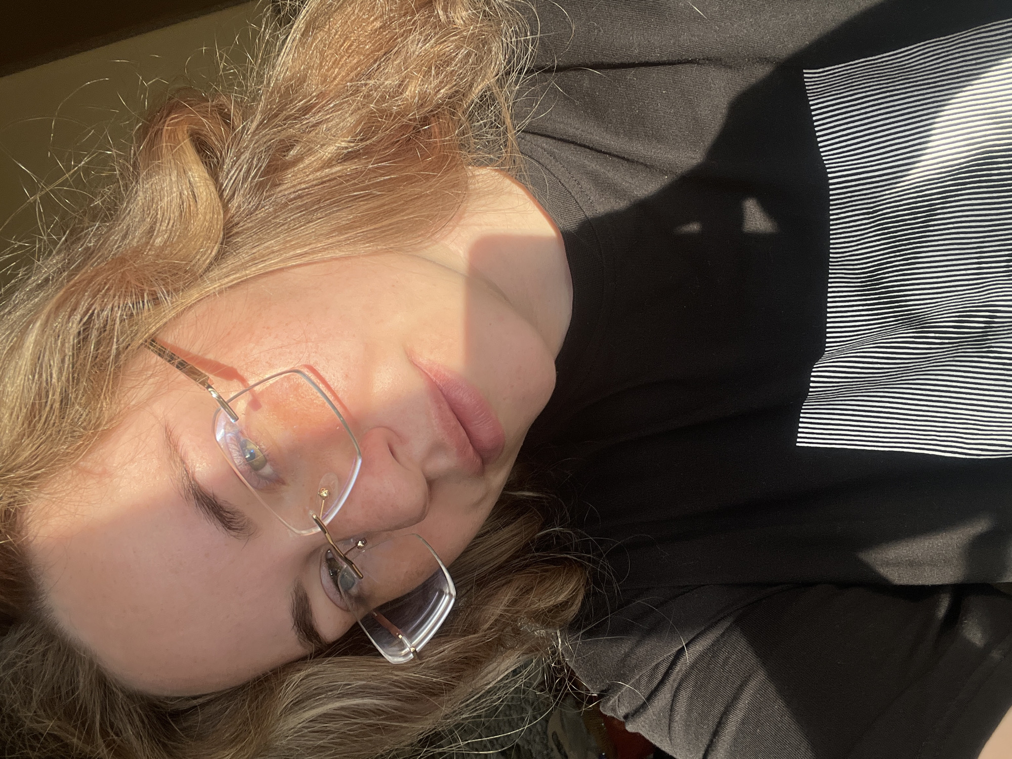 A woman with fair skin and light brown hair wearing glasses is oriented diagonally in the frame. She is wearing a black T-shirt with a horizontal white striped design on the upper right section. A strong, direct light source illuminates the left side of her face, creating harsh shadows. The background is blurry and indistinct. There is no visible indication that the image was taken in Сосново, Russia.