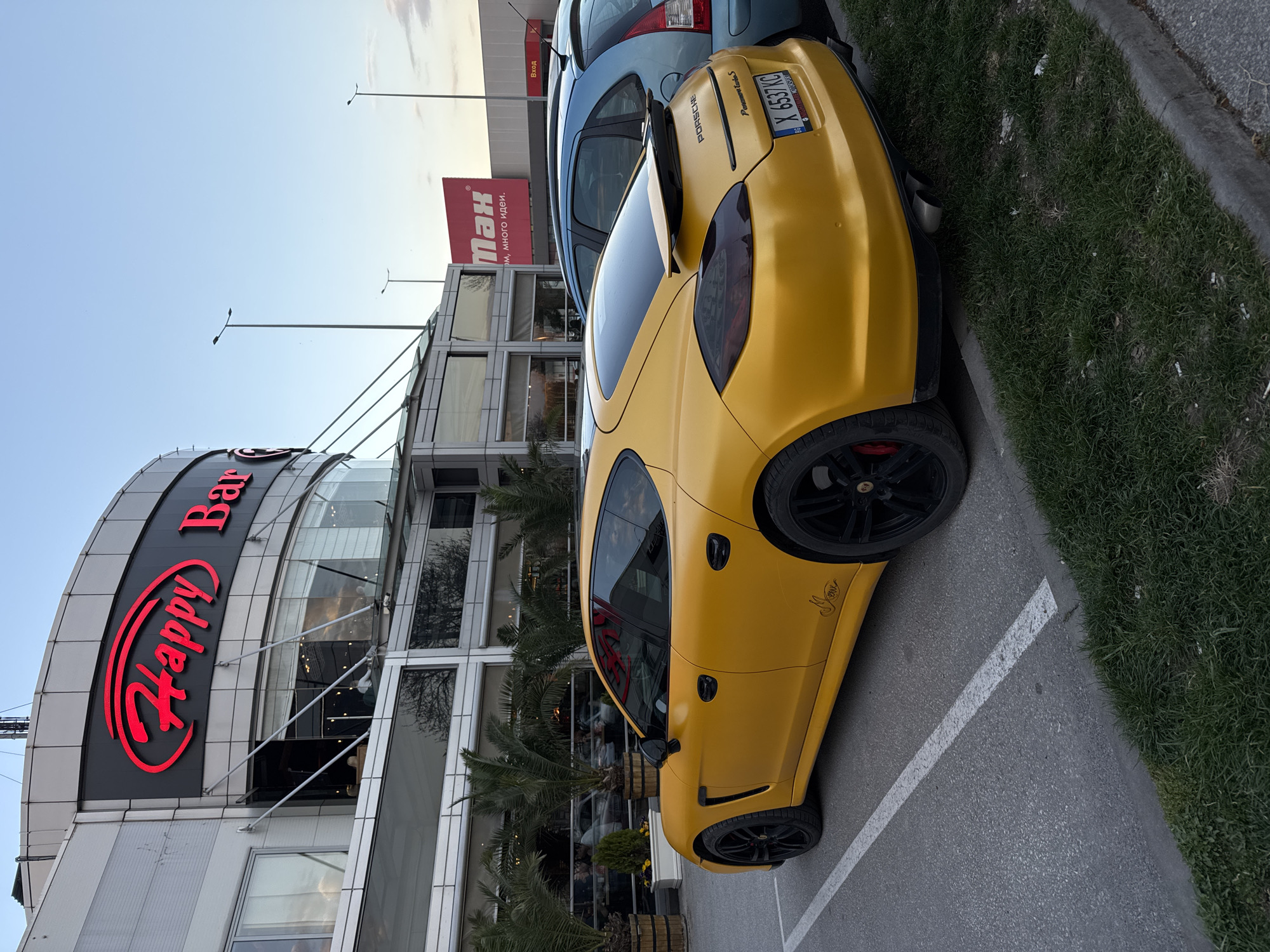 The image captures a yellow Porsche Panamera Twin S, distinguished by its license plate "X 6537KC", parked beside a blue car near the "Happy Bar & Grill" in Plovdiv, Bulgaria. The restaurant's prominent red neon sign is visible against its modern, multi-story building facade, which features glass panels and is flanked by palm trees. Another sign reads "MАХ", followed by Bulgarian text, situated near the blue car and the building. The scene is set in a parking lot bordered by a curb and a grassy area. The image also captures glimpses of the surrounding urban environment, including light poles and a partially visible sky, suggesting an outdoor setting during either dawn or dusk.