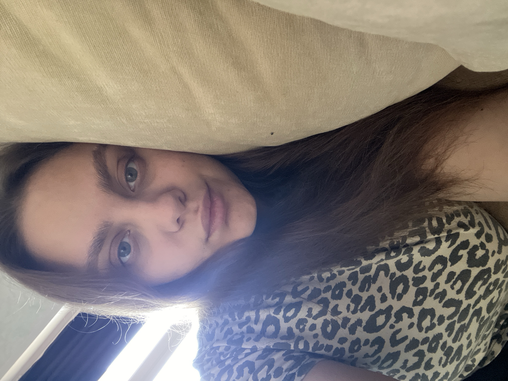 A woman with light skin, green eyes, and light brown hair is lying down. Part of a tan-colored pillow is behind her head. She is wearing a tan and black leopard print shirt. There is a bright light source coming from the left. The location in the city of Chaikovsky, Russia is not discernible from this image.