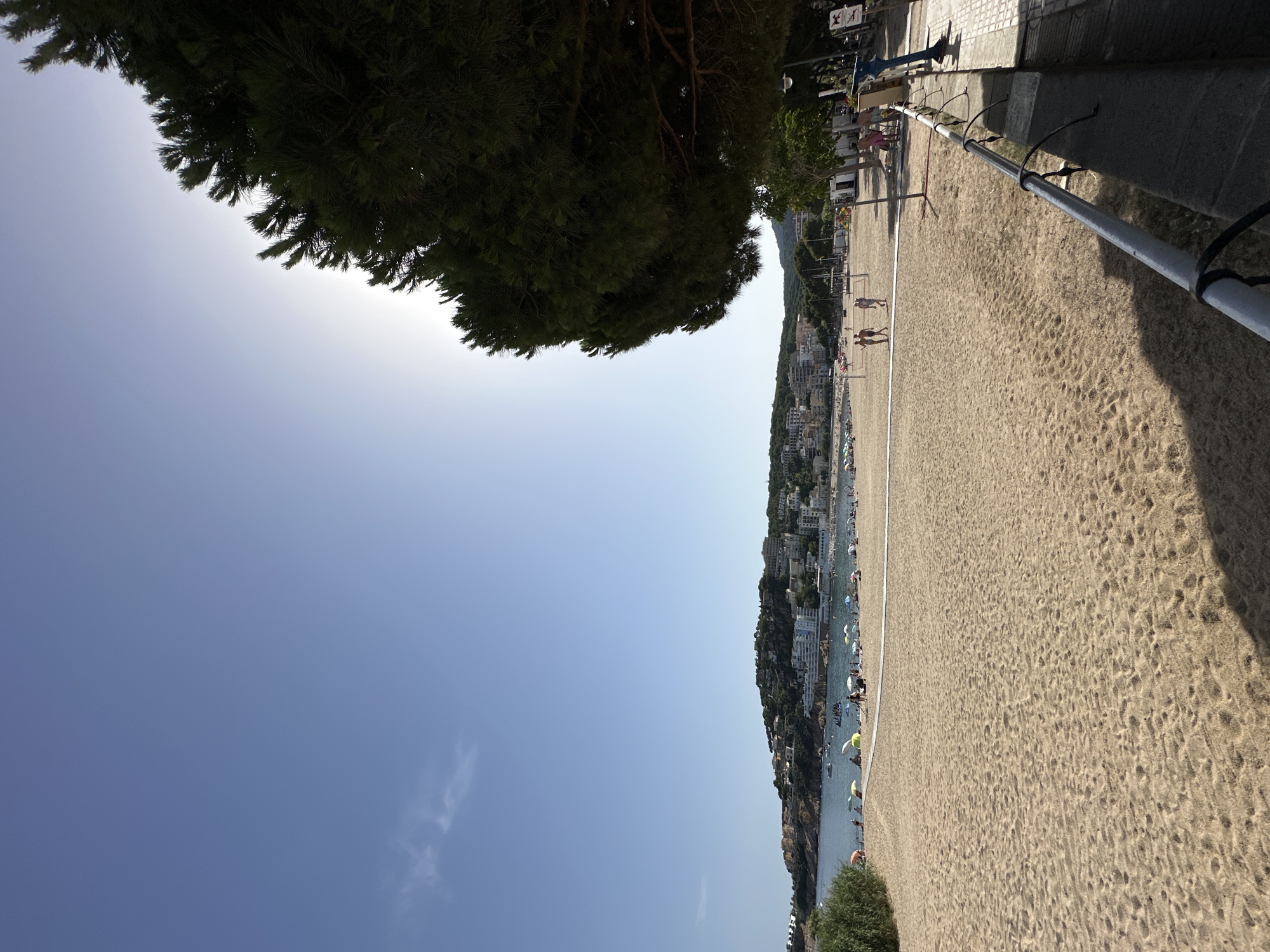This is a photograph taken at Platja Gran, the main beach in Sant Feliu De Guíxols, Spain, on a sunny day. The image is dominated by a wide expanse of golden sand in the foreground, showing footprints and scattered shadows. A white net, likely for a beach sport, stretches across the mid-ground of the sand.
Beyond the net, the beach continues towards the water, dotted with numerous colorful umbrellas and a scattering of beachgoers. The sea is calm and blue, with several small boats visible on the surface. In the background, a hillside is densely covered with buildings, hotels, and lush green vegetation, characteristic of the coastal town. A large pine tree on the right side of the frame partially obscures the bright sky and the sun, casting a shadow onto the foreground.
Several people are visible on the beach. Two individuals are walking towards the net in the mid-ground, and a group of people are standing or sitting near the water's edge. On the promenade to the right, there are more people, some appearing to be walking. A blue public telescope stands on the promenade, along with a metal railing. The overall impression is of a popular, vibrant beach destination.