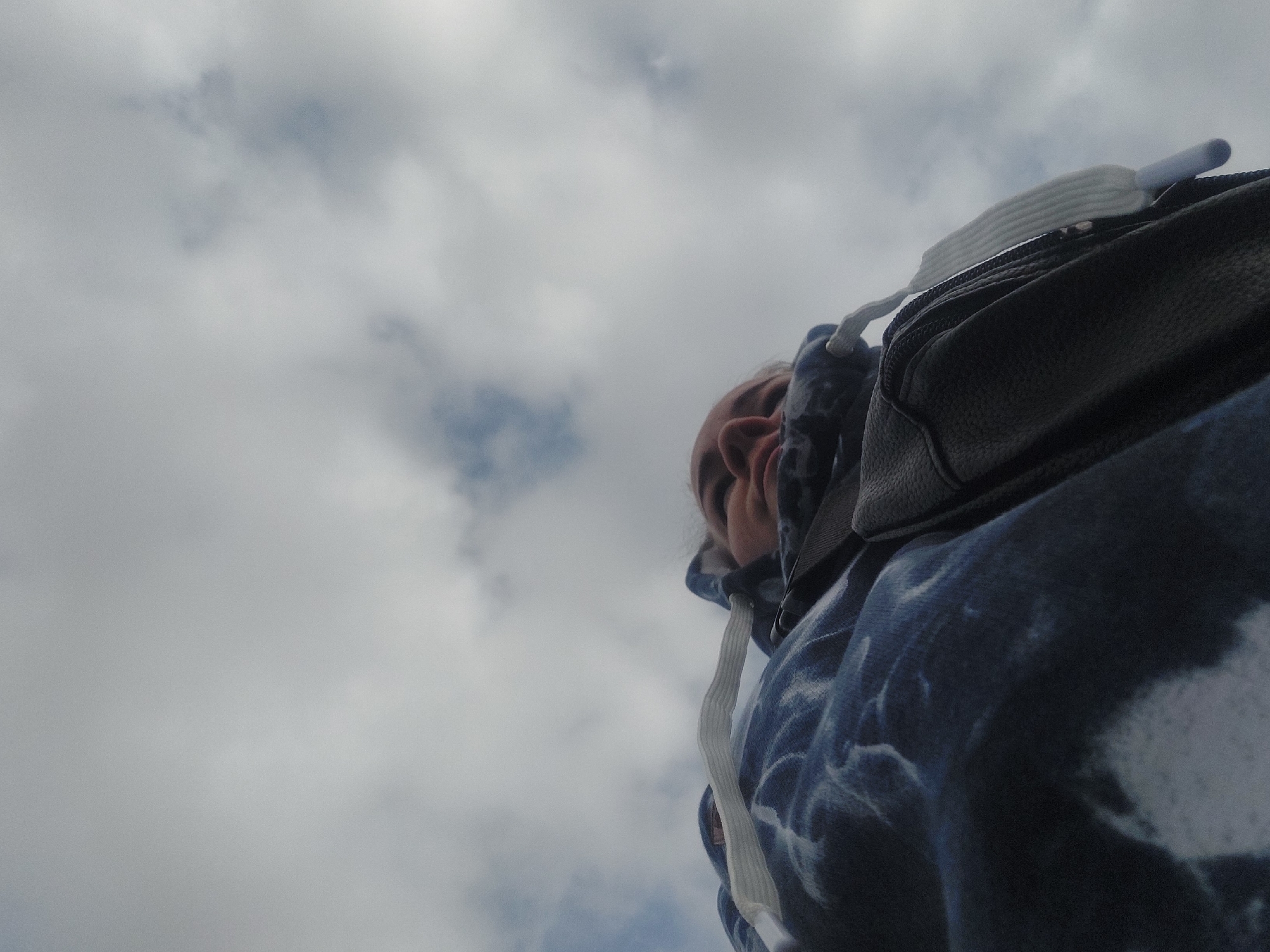 The image is a low-angle shot of a person looking up at a cloudy sky. The person's face is partially visible, and they are wearing a blue and white tie-dye hoodie. A black bag with a white strap is also visible. The background is filled with grey and white clouds. The location and specific details to confirm that this photograph was taken in Chaikovsky, Russia, are not apparent in the image. There are no visual cues to confirm a specific location.