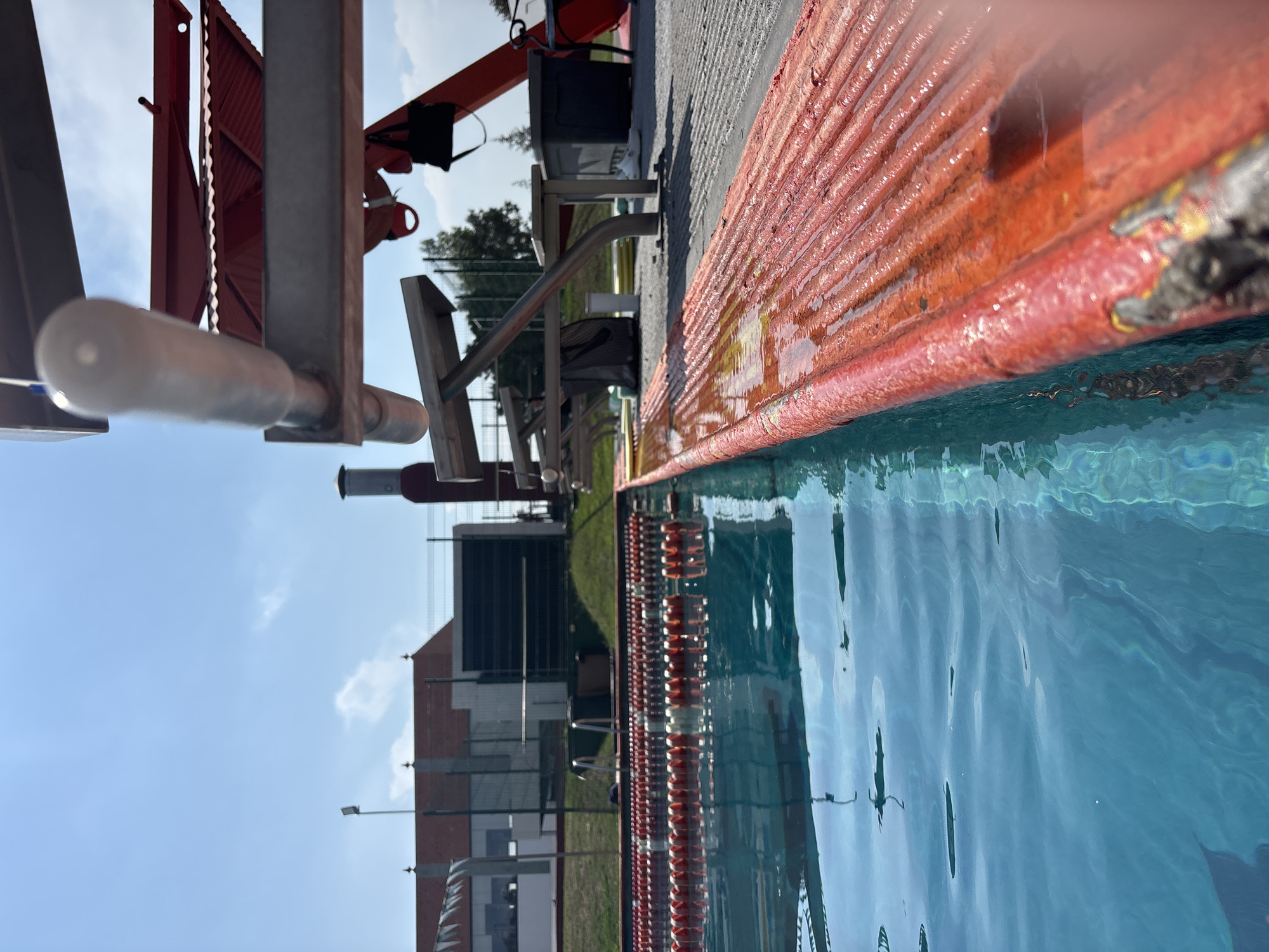 The image depicts a swimming pool at the Unidad Deportiva "San Bernardino" in San Andrés Cholula, Mexico. The pool water is blue and rippled. Orange-and-white lane dividers run along the length of the pool. A red metal platform borders the pool, with a diving platform and starting blocks visible. In the background, there is a brick building with black window frames, green grass, and a blue sky with some clouds.