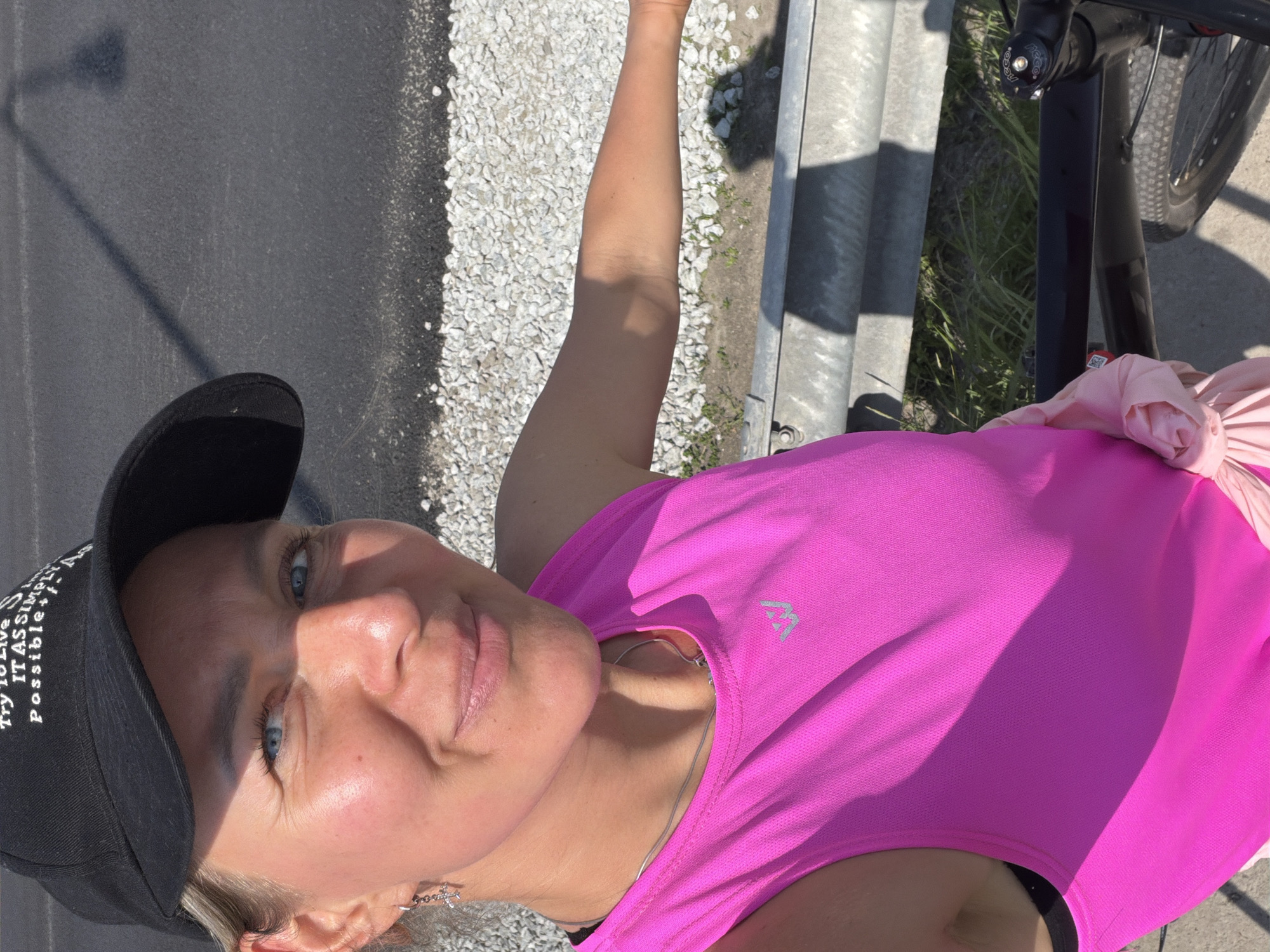 A woman is taking a selfie outdoors, with a black baseball cap on her head and a bright pink tank top. She has blue eyes and light skin. Her left arm is extended, and she is holding a light pink cloth. A bicycle is partially visible in the foreground. In the background, there is an asphalt road and a metal guardrail. The scene appears to be near a road in Chaikovsky, Russia.