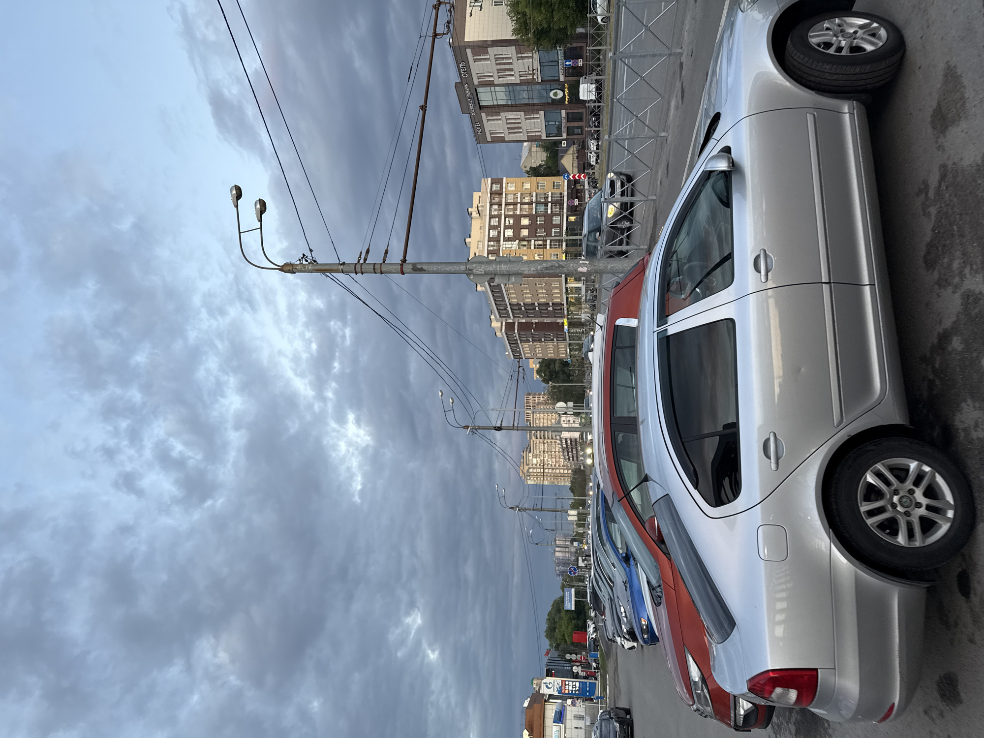 The image captures a street scene in Kazan, Russia, likely during the late afternoon or early evening, given the overcast sky and muted lighting. In the foreground, a silver sedan is parked. Behind it, a row of other cars is visible, including an orange car and a blue car. The cars are parked along a street lined with a metal fence. In the background, there are several multi-story buildings, some appearing to be residential complexes and others commercial, including one labeled "HOTEL". Streetlights are prominent, with one double-headed streetlight visible closer to the camera. Utility poles and wires crisscross the sky, typical of urban infrastructure. The overall atmosphere is one of a somewhat cloudy day in an urban environment.