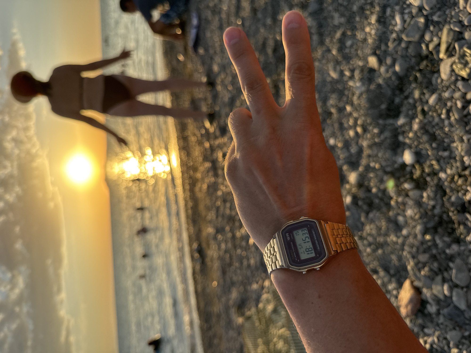 The image depicts a close-up of a person's wrist wearing a gold digital Casio watch, displaying the time 18:15:33. The watch is on the left wrist, and the person's hand is extended forward, making a peace sign with their index and middle fingers. The background is a blurred scene of a beach at sunset. The sun is visible low on the horizon, casting a golden glow on the water and the sky. Several people are visible in the water and on the shore, appearing as indistinct figures due to the blur. The ground in the foreground consists of pebbles. The setting is a beach, likely in Zharbulak, Kazakhstan, given the prompt, though the image itself does not provide explicit geographical markers.