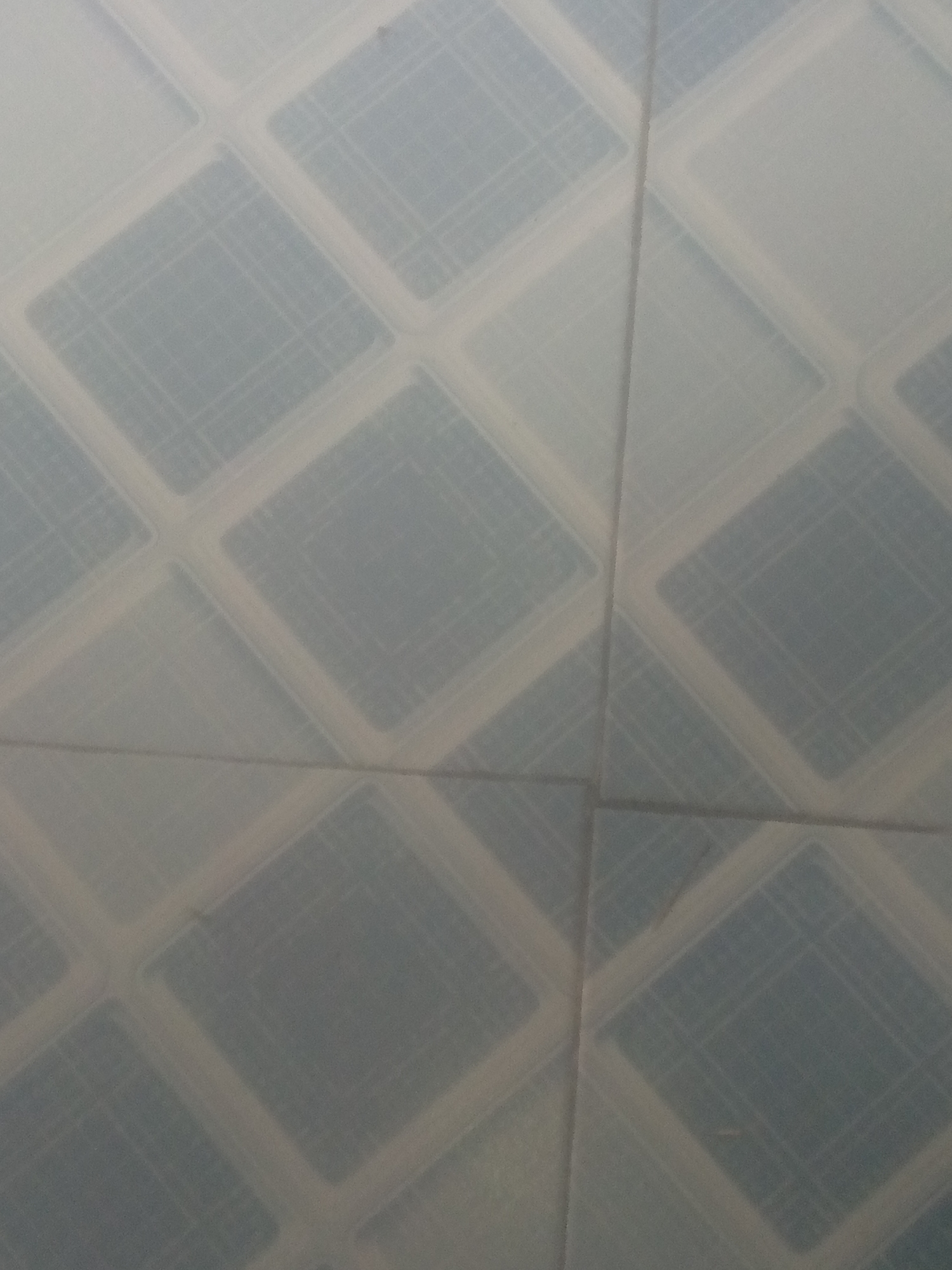 The image is a close-up, low-angle shot of light blue patterned tiles, likely on a wall or floor. The pattern consists of interlocking diamond shapes outlined in a lighter, off-white color. Within each diamond, there is a subtle grid-like pattern of fine lines. The lighting is diffused, creating a soft, somewhat hazy appearance. There are no people or other discernible objects in the frame, and the scene appears to be an interior space. The location is Unknown, Unknown.