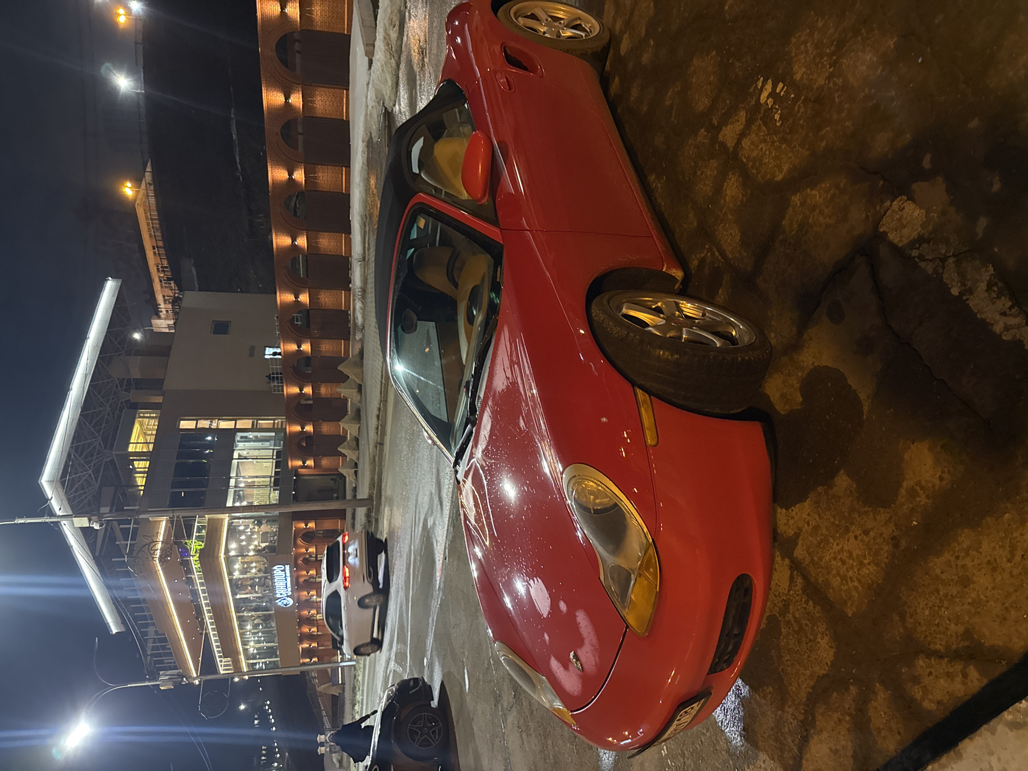 The image shows a red convertible car parked on a wet street in what appears to be an urban environment at night. In the background, there is a multi-story building with illuminated windows, likely a commercial or entertainment establishment. There are other cars parked on the street, and possibly pedestrians nearby, though details are limited. The surroundings appear to be urban, with street lights and modern architecture. The scene is located in the city of Izhevsk, Russia, specifically near the "Gambrinus" restaurant, identified by its signage on the building.