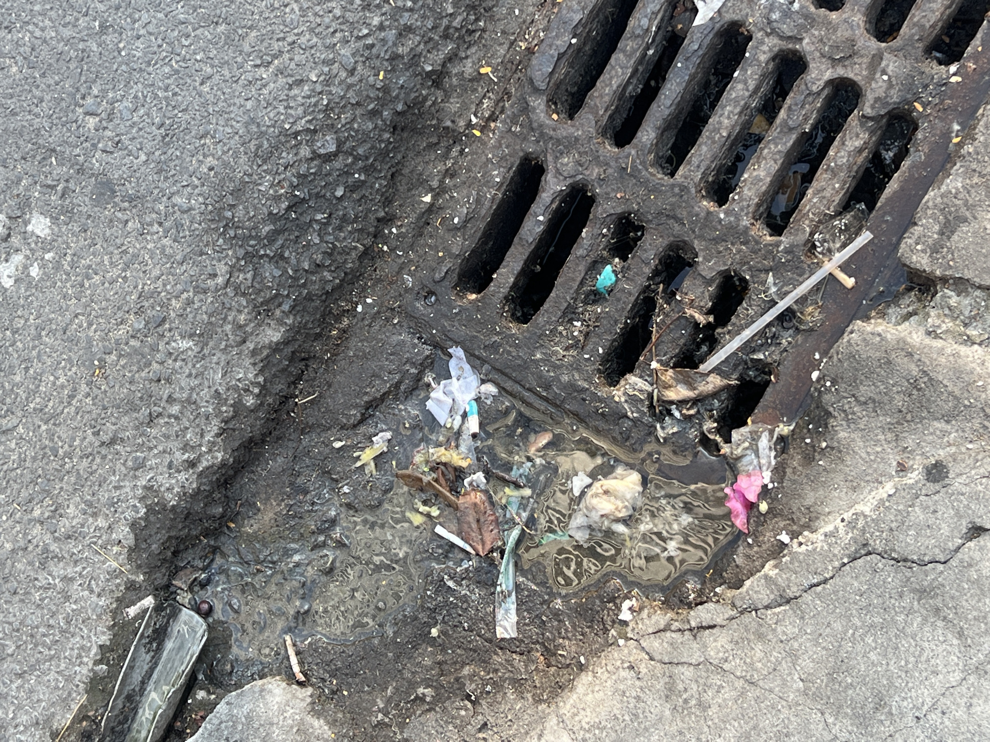 The image depicts a storm drain in Nha Trang, Vietnam, heavily clogged with litter and debris. The drain is constructed of metal grating and is partially obscured by a pool of stagnant water filled with paper, plastic fragments, leaves, and discarded items. The drain is situated between asphalt pavement and cracked concrete.