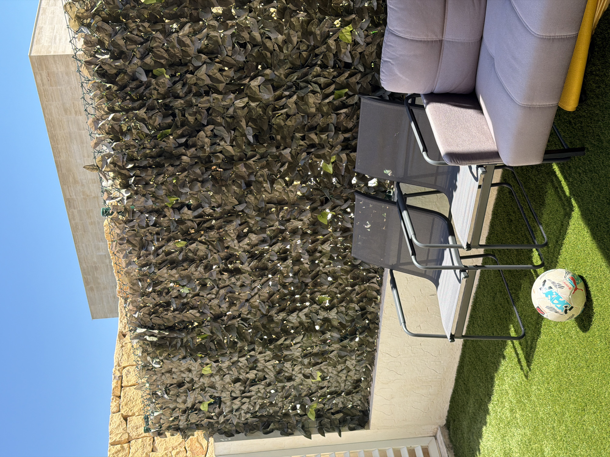 In Finestrat, Spain, a sunny outdoor patio features artificial grass. Two dark gray metal chairs with mesh seats and backs are positioned on the grass. To the right, a light pink upholstered sofa with black metal legs is visible. A multicolored soccer ball rests on the grass in front of the chairs. Behind the furniture, a dense hedge of artificial dark green leaves forms a backdrop, secured by green netting attached to a textured beige wall. Above the hedge, a light beige stone wall topped with a light beige building can be seen against a clear blue sky.