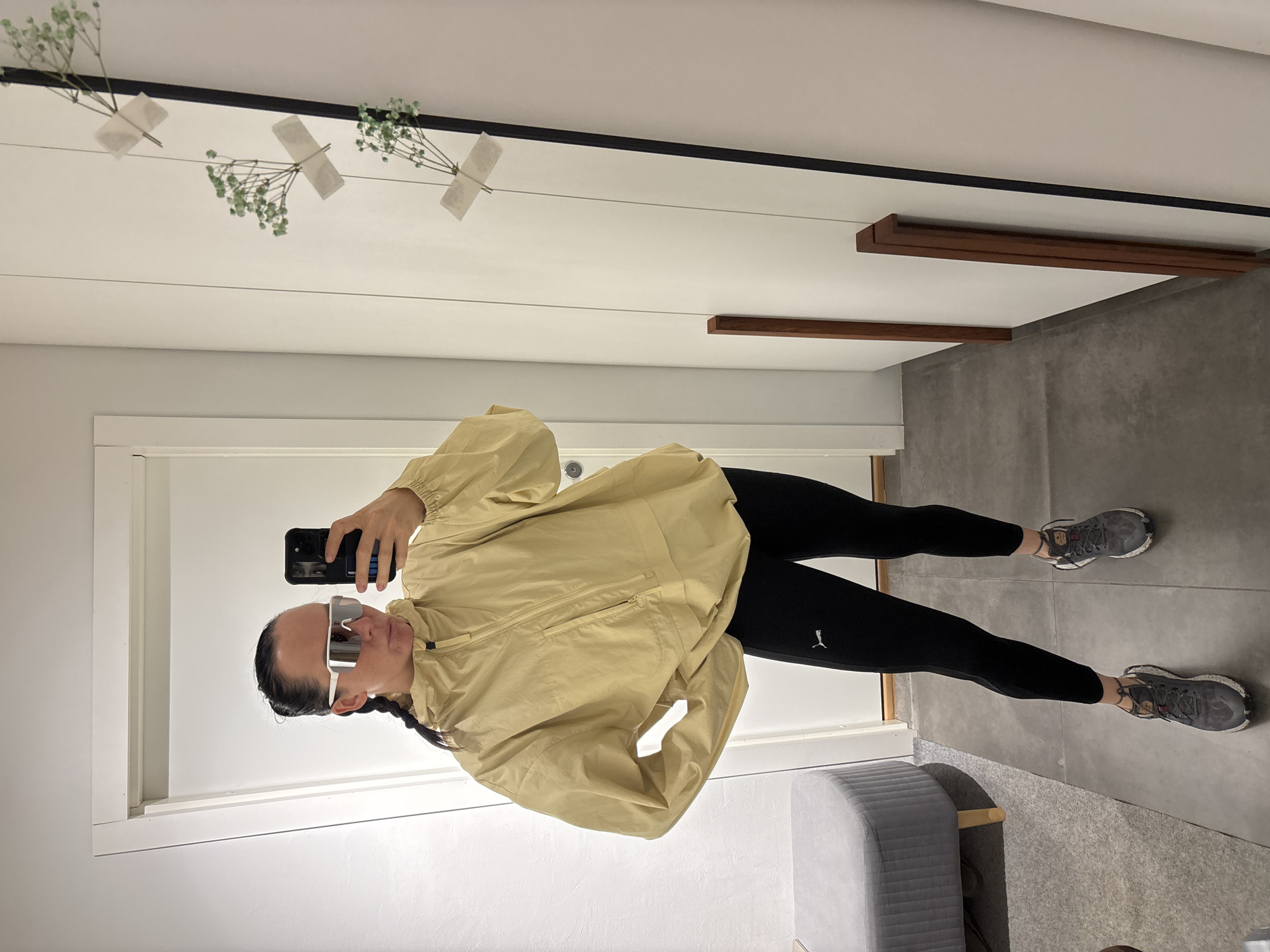 A person is captured taking a mirror selfie indoors in Chaikovsky, Russia. The individual, likely a woman, has dark hair pulled back in a braid and is wearing large, futuristic-style white and silver reflective sunglasses. She holds a black smartphone to take the picture, with her arm visible.
Her outfit consists of a light beige or tan anorak-style jacket with a zipper and pockets, paired with black athletic leggings that feature a white "Puma" logo on the left thigh. She wears grey athletic shoes with visible wear on the soles.
The setting appears to be a modern indoor space, possibly a hallway or room. The mirror reflects white walls, accented by a thin black horizontal strip near the top. Decoratively taped to a string or wire reflected across the upper part of the mirror are several small sprigs of green baby's breath flowers. Further to the right in the reflection, dark brown wooden shelves or decorative elements are visible against a lighter wall. The floor is composed of large grey tiles, and a grey upholstered bench or ottoman is partially visible in the lower-left foreground of the image. The overall atmosphere is casual and contemporary.
