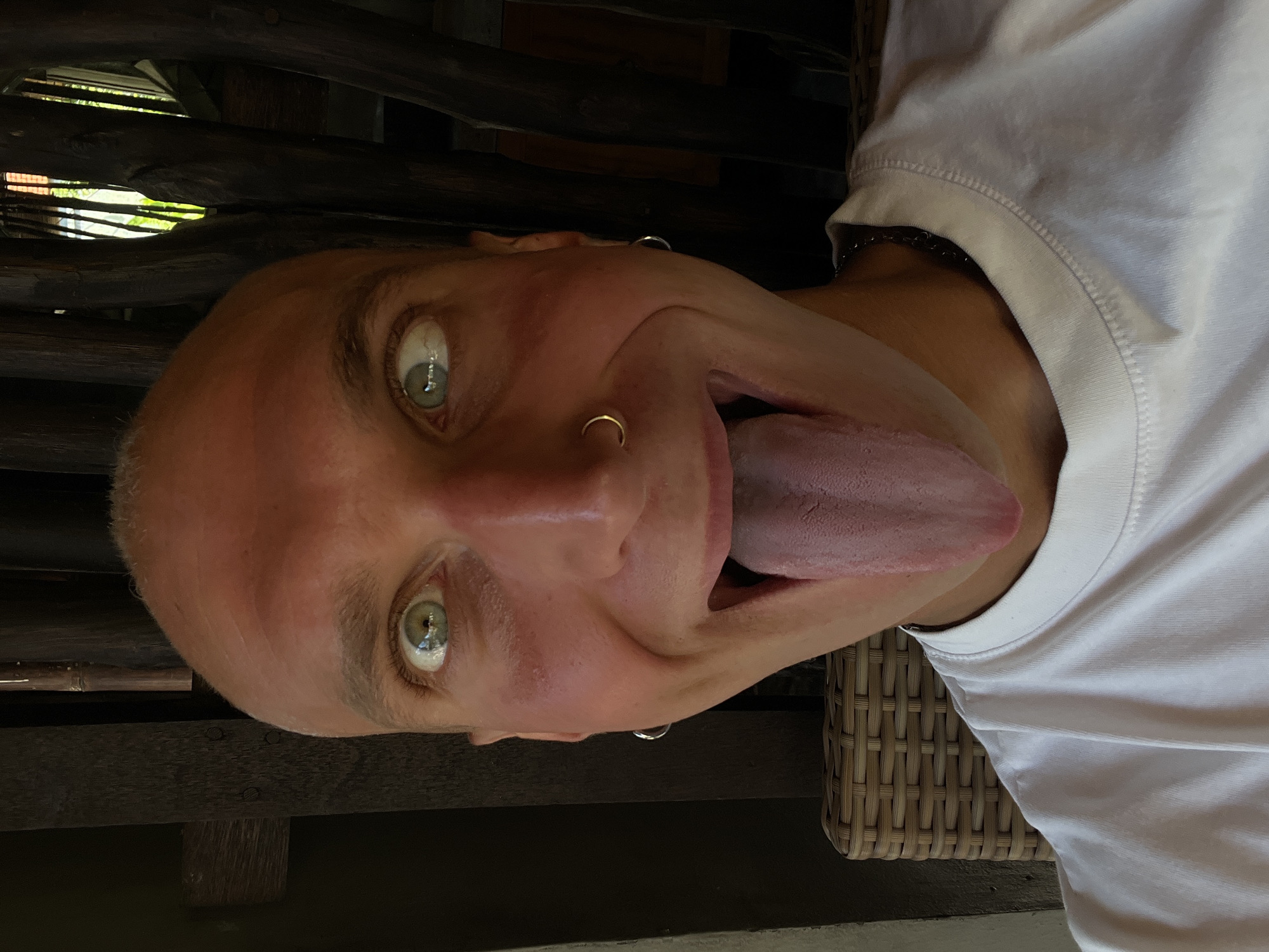 A close-up selfie shows a bald, fair-skinned person with light green eyes sticking out their long, pink tongue. They are wearing a white t-shirt and a silver chain necklace. A gold hoop is visible in their left nostril, and a silver hoop earring is in their left ear. Their eyes are looking in different directions, giving a cross-eyed appearance. The background is out of focus, but appears to be an outdoor setting with dark wooden pillars and some greenery visible through them. The person is seated on a wicker chair. The image was taken in the city of Mengwi, Indonesia.