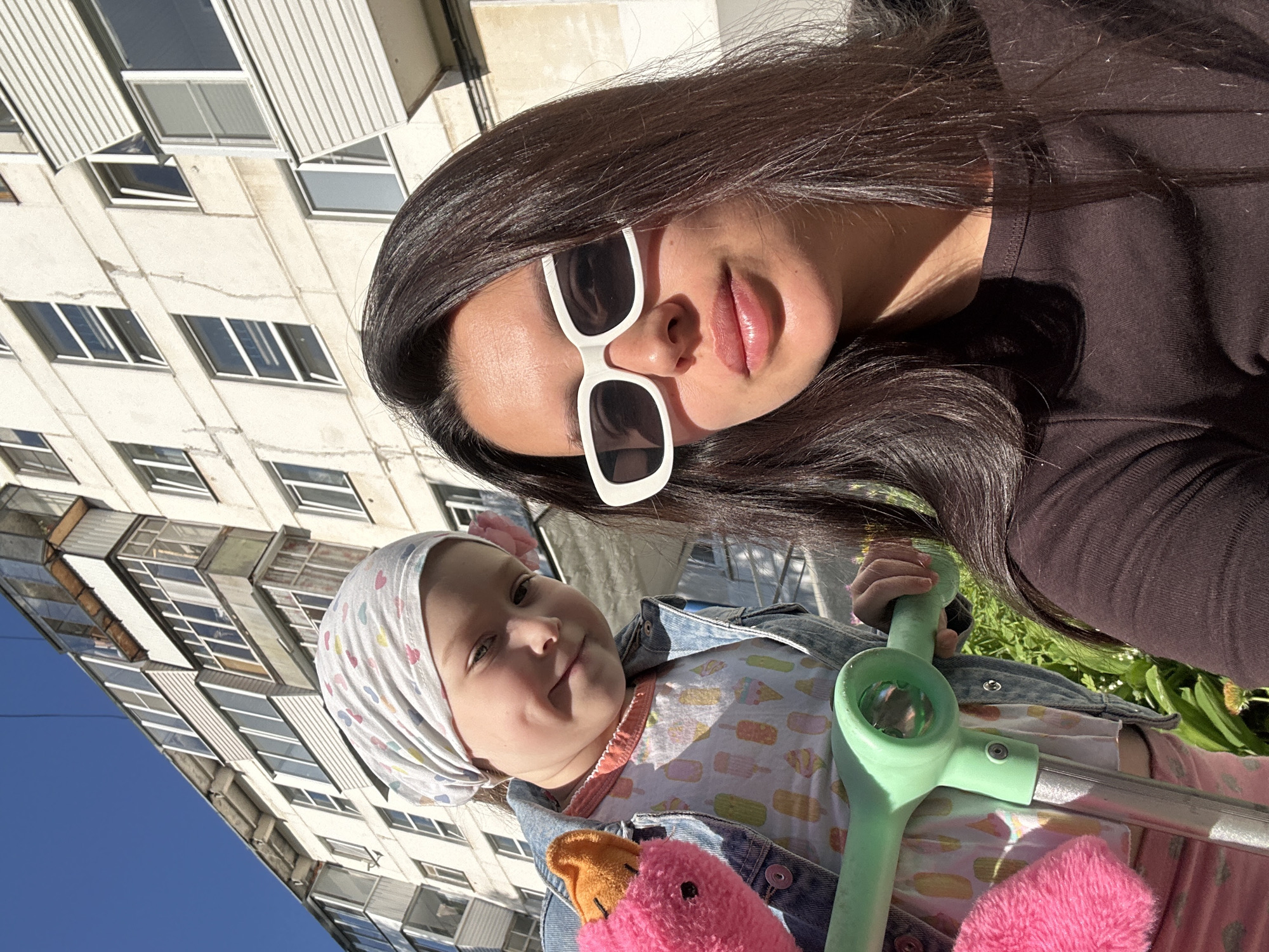 A selfie captures a woman and a child in front of an apartment building. The woman wears sunglasses and a dark top. The child, sporting a patterned headscarf, sits in what appears to be a stroller or riding toy with pink and orange plush accessories. The building has multiple windows. Based on the photographer's reported location, this is likely a scene in Chaikovsky, Russia.