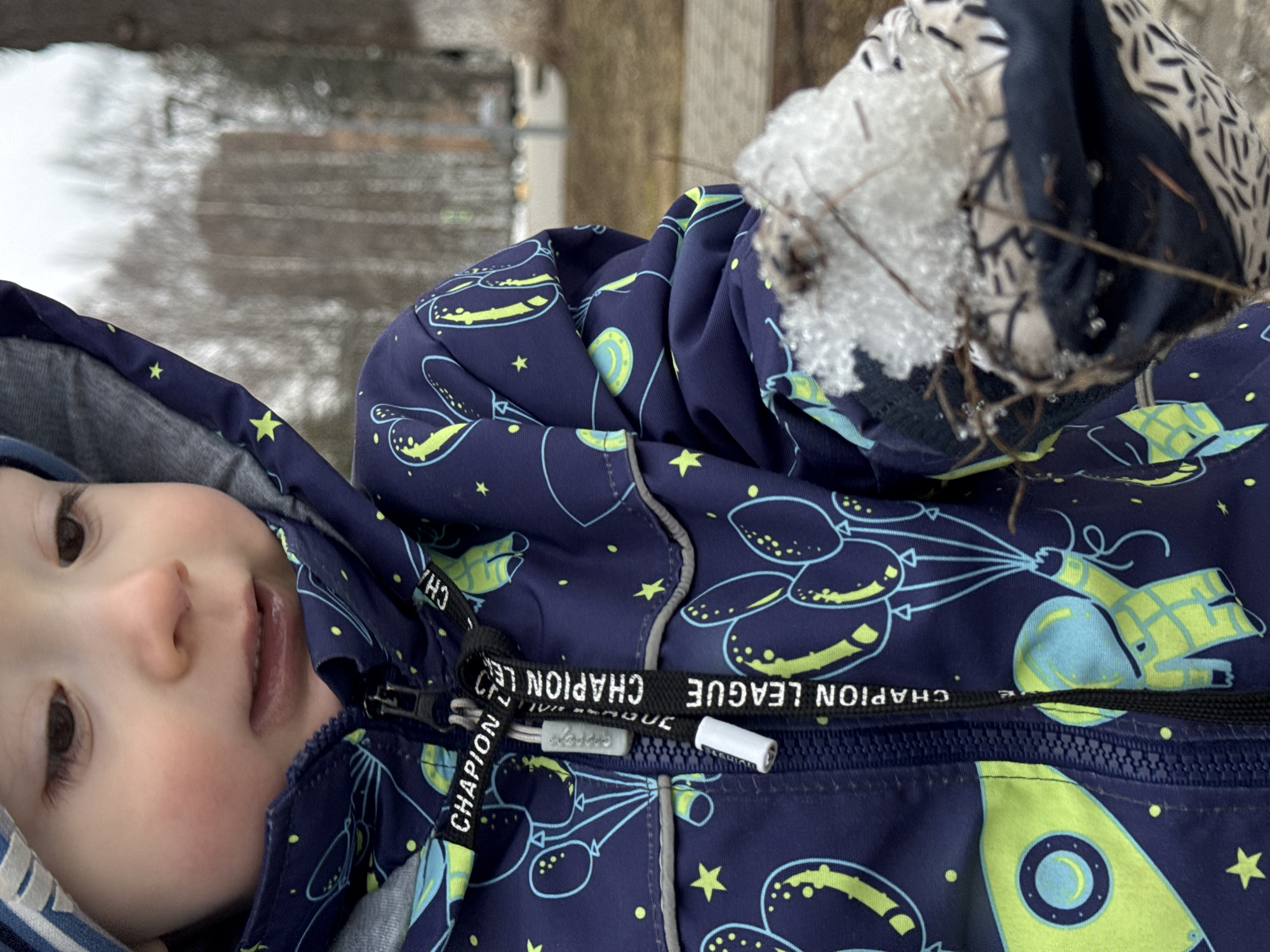 A close-up, slightly angled shot shows a young child wearing a dark blue hooded winter jacket with a pattern of balloons, astronauts, and stars. The jacket is zipped up, revealing the word "CHAPION LEAGUE" on the zipper pull. The child's face is visible with brown eyes, rosy cheeks, and a slightly open mouth. Snow and twigs are visible near the top edge of the image, resting on the jacket. The background is blurred and appears to show a wooden structure and an outdoor environment, possibly with remnants of snow. The location is not possible to be determined.