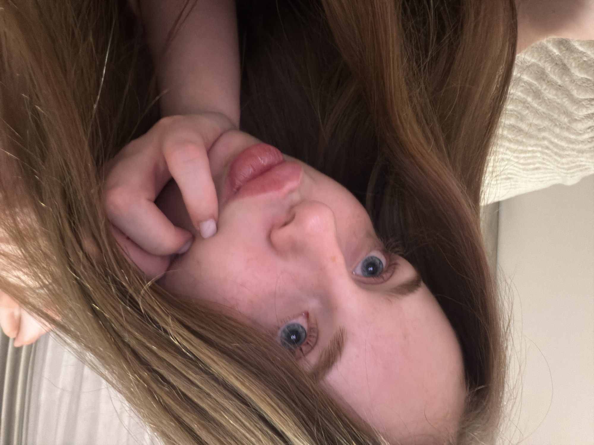 The image presents a close-up, angled indoor portrait of a young female, situated within an unspecified room in Saint Petersburg, Russia. The female exhibits fair skin, light blue eyes, and long, straight, light brown hair that frames her face and flows around her head. She is positioned as if lying down or reclining, with her head slightly tilted, making direct eye contact with the camera. Her lips are pursed in a 'duck face' expression. Her right hand is visible, with fingers curled and light-colored nail polish, supporting her lower cheek/chin. The background, softly out of focus, reveals a light-colored, wavy-textured fabric, likely a blanket or bedspread, on the left. To the right, partially obscured, are light-colored, vertically pleated curtains or drapes. The ambient lighting appears soft and uniform.