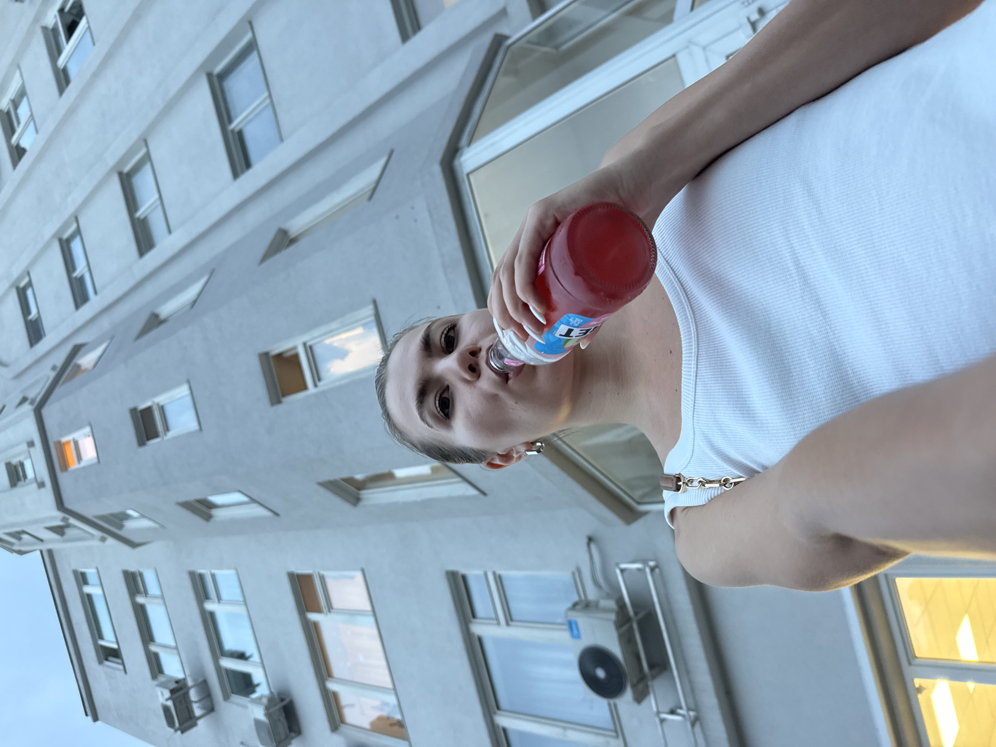An outdoor, low-angle, medium shot captures a young woman from the chest up, holding a bottle of red liquid to her lips and drinking from it, as she stands before a light gray, multi-story building. The woman is positioned in the lower-right quadrant of the frame, her left shoulder and arm extended to the left, holding the bottle with her right hand. She is wearing a white ribbed tank top and a silver hoop earring in her left ear. Her blonde hair is pulled back in a ponytail, and her eyes are directed towards the camera. The building behind her, shot from a low angle, extends upwards and to the left, with multiple windows visible. The sky above is overcast and blue. An air conditioning unit is mounted on the exterior wall to the left of the woman. The overall scene is likely an urban environment in the city of Unknown, Unknown.