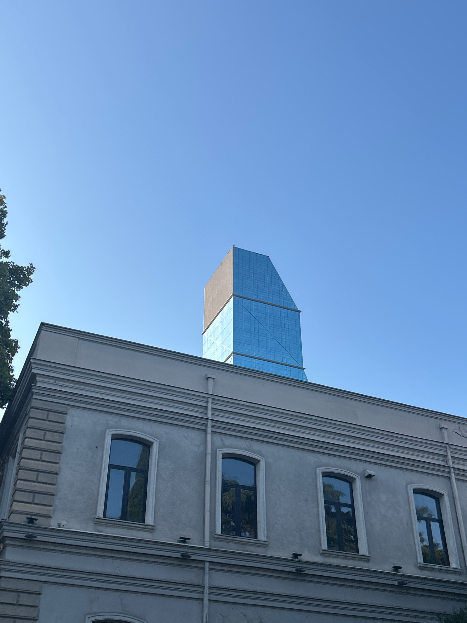 The image shows a clear blue sky above a building in Tbilisi, Georgia. In the foreground, a grey, classical-style building with decorative molding and arched windows is visible. To the right of this building, a modern skyscraper with a blue, glass facade rises into the sky. The skyscraper has a distinctive angular design. A tree with dark green foliage is partially visible on the left edge of the frame. The scene appears to be an outdoor view of urban architecture.