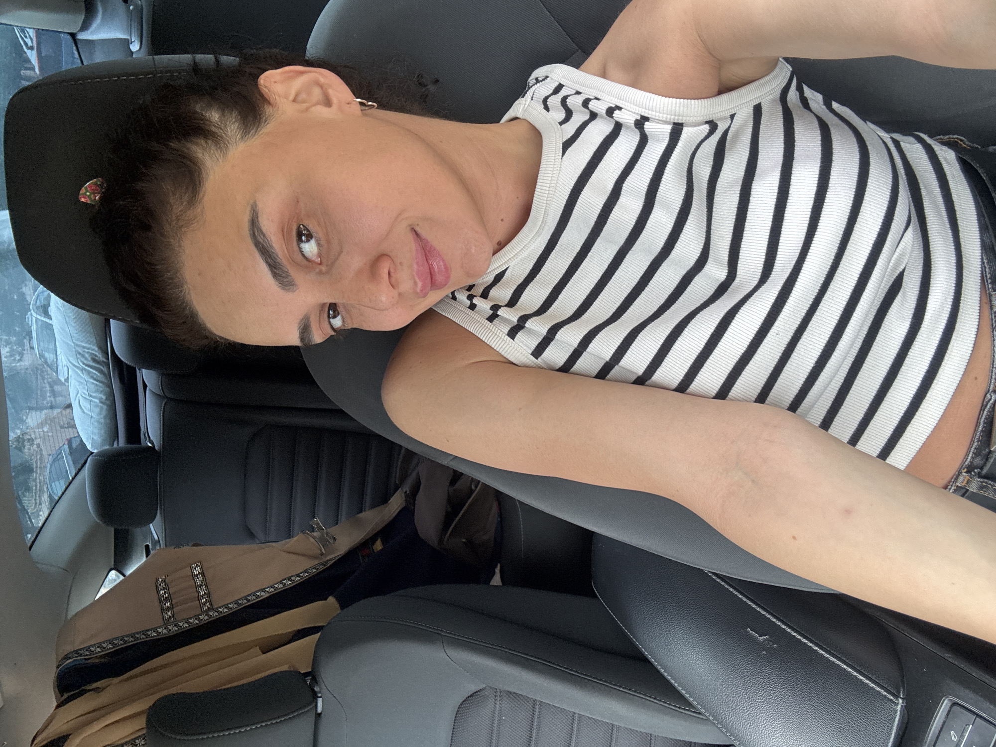The image shows a woman sitting in the driver's seat of a car. She is wearing a black and white striped tank top and dark denim jeans. Her hair is dark brown and pulled back in a ponytail. She is looking at the camera and smiling. In the background, there is a beige jacket hanging on the back seat. The interior of the car appears to be black leather. The exterior of the car is not visible, but the background outside the car shows buildings and trees, suggesting an urban environment. The specific location in Tbilisi, Georgia cannot be determined from the visual information provided.