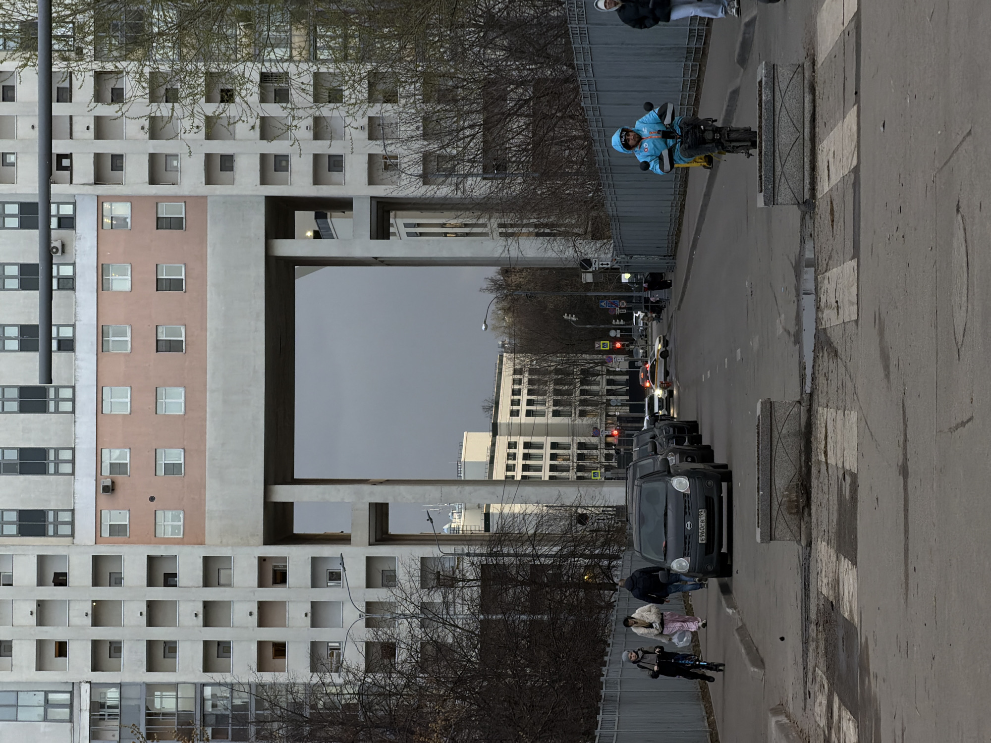 The image shows a street scene in Khimki, Russia, featuring a large concrete building with multiple open-air balconies on the left side of the frame. The building has a central section that appears to be a lighter pink color. A concrete structure spans above the street, creating a visual frame.
A line of cars is parked along the right side of the street, with a pedestrian crossing visible. Several people are walking along the sidewalk, and one person is riding a bicycle on the street.
The sky is overcast. The overall environment seems urban and functional, typical of a residential district.