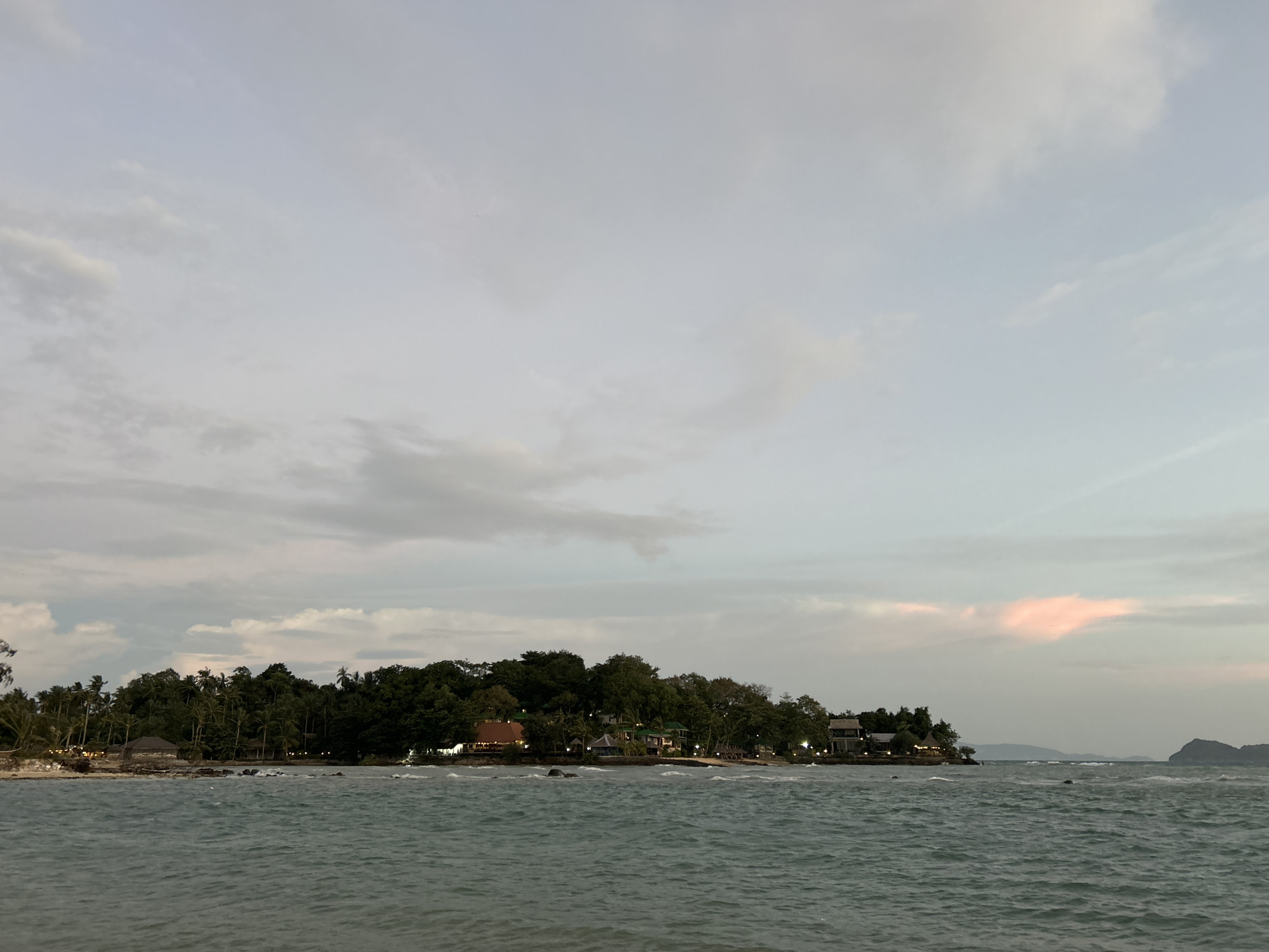 The image shows a tropical coastline at dusk in the Ko Pha-ngan District of Thailand. The foreground is dominated by the choppy turquoise sea, with gentle waves breaking on a sandy shore in the distance. Along the shoreline, a dense line of palm trees and other tropical vegetation separates the water from a cluster of buildings. These buildings appear to be resort accommodations, with visible lights suggesting that people are present, although no individuals are discernible in the frame. The architecture suggests a relaxed, vacation-oriented setting, with some structures featuring thatched roofs and open-air designs. The sky above is a muted blend of grey and pale blue, with scattered clouds, some tinged with pink from the setting sun, hinting at the fading light. In the far distance, a silhouette of a larger island or mountain range is visible on the horizon. The overall atmosphere is tranquil and serene, characteristic of a tropical evening.