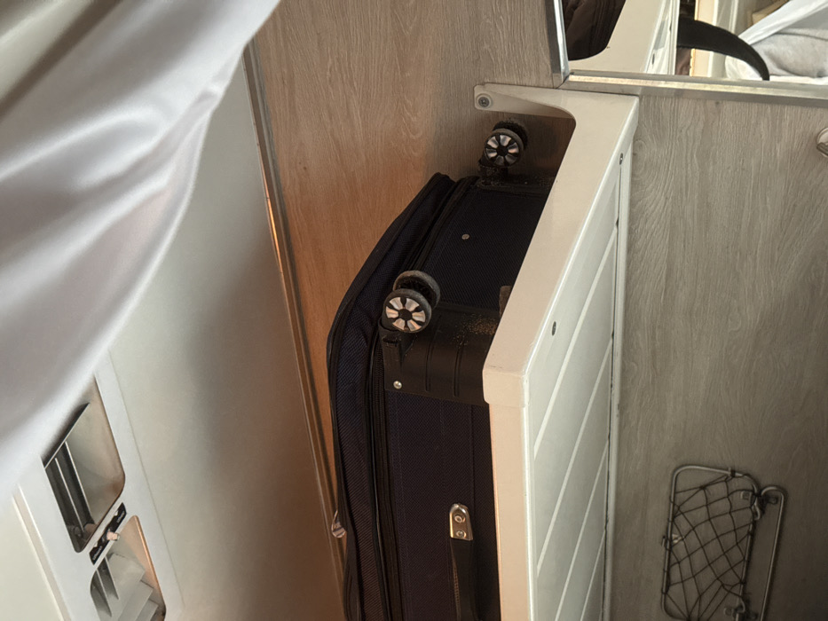 In Gryazi, Russia, an interior scene is depicted focusing on a dark blue, soft-sided wheeled suitcase resting on a white, ribbed overhead shelf. The suitcase, positioned horizontally, displays two of its four wheels, which exhibit a distinctive star-like spoke pattern and visible accumulation of dirt or grime. The white shelf itself is mounted to a light wood-grain patterned wall, secured by a white L-bracket on the right. Below the shelf, a small, black metal mesh storage rack is affixed to the wood-grain wall. Above the suitcase and shelf, a light-colored (likely white) wall or ceiling features two rectangular vents with metallic grilles in the upper left corner. A white, draped fabric, consistent with a curtain, is visible along the right edge and upper right portion of the image. To the far right, a reflective surface, possibly a mirror, partially reveals indistinct dark and light objects. The overall environment suggests a compact travel compartment, such as a train cabin or a recreational vehicle. No individuals are visible.