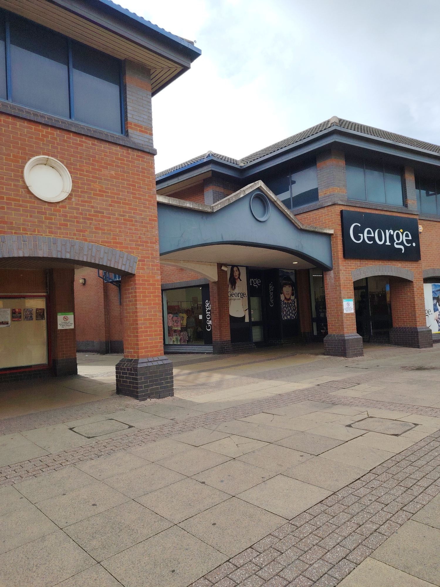 The image captures the exterior of a retail building, identified as a George clothing store, situated in Peterborough, United Kingdom. The scene is depicted under an overcast sky.
The primary structure is a multi-story building constructed predominantly with reddish-brown brick, featuring dark grey brick detailing at its base and window sills. It is topped with a dark grey tiled roof exhibiting an overhang. Large, dark, reflective windows are present across the upper levels. A prominent black sign with white lettering stating "George." is mounted on the right-hand facade. The main store entrance is recessed, forming a covered walkway supported by brick pillars. Large display windows within this recess showcase clothing and mannequins, with additional internal "George" and "George.com" branding visible. A dark grey, circular architectural detail is centered above the recessed entrance.
To the immediate left, a portion of an adjacent brick structure is visible, featuring an arched opening at ground level and a white circular fixture on its upper wall.
The ground surface in the foreground is paved with large, light grey square slabs, interspersed with sections of reddish-brown block paving. Several rectangular utility access covers are integrated into the paved surface. No individuals are present in the scene.