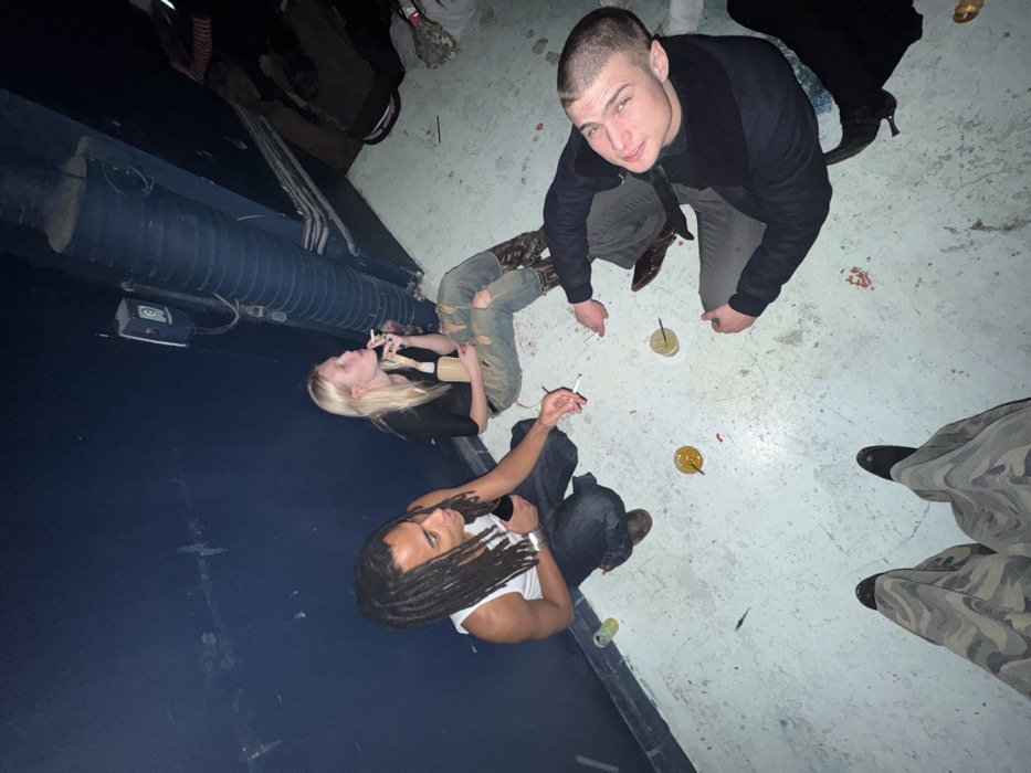 An indoor, dimly lit scene captures four individuals gathered on a worn, light-grey concrete floor. The precise location within Paris, France, is not visually identifiable from the image.
Three individuals are seated on the floor against a dark blue or black wall. To the left, a male with dark skin and dreadlocks wears a white tank top and dark jeans. He holds a cigarette in his right hand and a dark object, possibly a phone, in his left, looking towards the right. Adjacent to him, a female with long blonde hair wears a black top and ripped light-wash jeans, sitting with her legs extended. She holds a lit cigarette to her mouth with her right hand and a light-colored bottle, possibly champagne, in her left, looking upward. She wears dark, pointed-toe boots. To the right, a male with light skin and a short buzz-cut wears a dark long-sleeved shirt and dark pants, facing the camera with a slight smile. He also wears dark, pointed-toe boots.
The foreground shows the legs and feet of a fourth individual, wearing camouflage-patterned wide-leg pants and dark shoes, indicating the camera perspective is from a standing or crouching position.
On the floor, three clear plastic cups containing an amber-colored liquid are present, along with a small green cup near the first male. Scattered red and white debris, possibly confetti or cigarette ash, and several cigarette butts are visible on the floor. An electrical box or light switch is mounted on the dark wall to the upper right of the first male, and a large, dark, corrugated pipe runs diagonally upwards along the right side of the frame, indicating an industrial or utility space. The ambient lighting is dim.