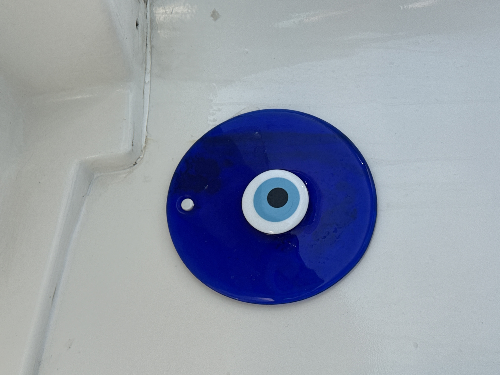 The image shows a close-up view of a blue evil eye talisman hanging on a white, textured surface. The talisman is circular with a raised concentric design in the center, featuring black, light blue, and white circles. A small white loop for hanging is visible at the top of the blue disc. The white surface appears to be part of a building's exterior or interior, possibly a wall or a boat hull, given the molded lines and slightly uneven texture. The lighting suggests an outdoor or well-lit indoor environment. There are no people or any other objects clearly visible in the frame.