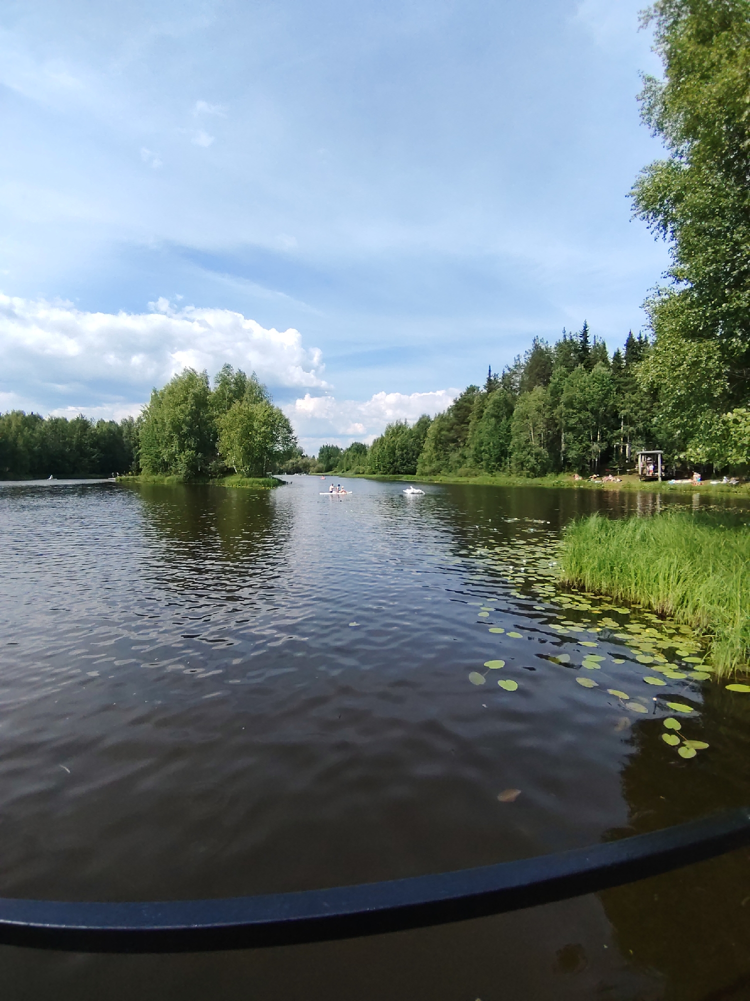 In the city of Vyltydor, Russia, a serene lake scene unfolds under a partially cloudy sky. The foreground features a dark body of water with gentle ripples, reflecting the sky and the surrounding greenery. Lily pads are scattered across the surface of the water, particularly on the right side of the frame.
A small, tree-covered island is visible in the middle distance, adding a natural focal point to the lake. On the water, two small kayaks with people can be seen paddling, suggesting recreational activity. Further in the background, the shoreline is densely lined with various trees, creating a lush, green backdrop.
On the right side of the lake, a wooden structure resembling a small gazebo or covered seating area is present, with a few individuals relaxing nearby. The overall atmosphere is one of peaceful outdoor recreation in a natural setting.