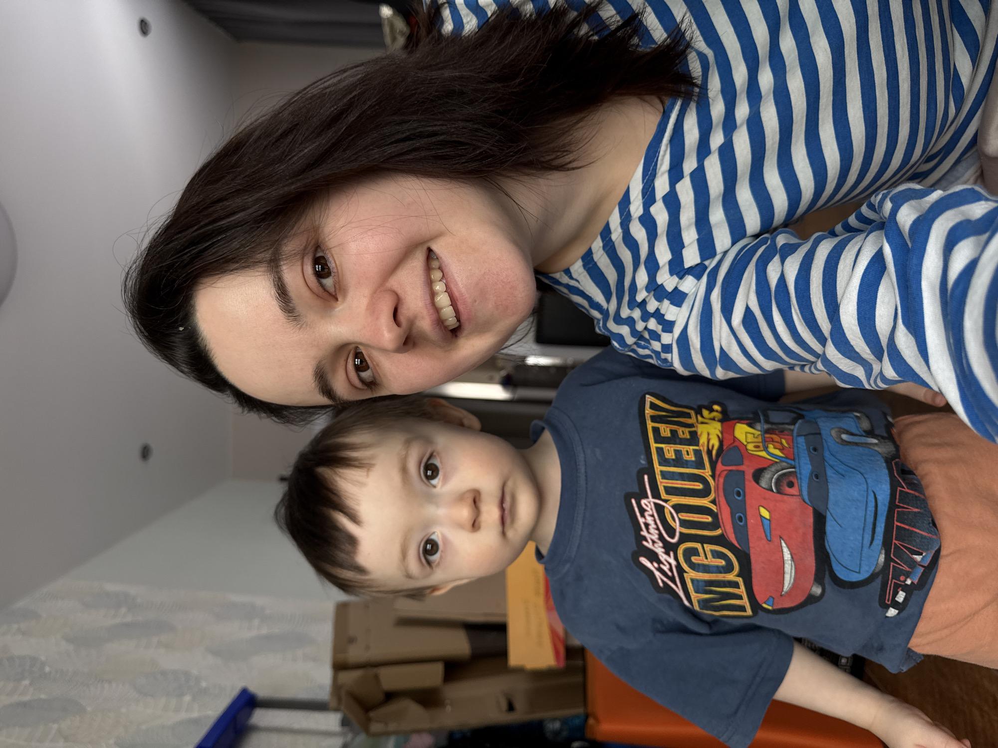 A woman and a young child are posing for a selfie in what appears to be an indoor environment. The woman, positioned at the top of the frame, has dark hair and is wearing a blue and white striped shirt. She is smiling broadly. Below her, the child is wearing an orange pants and a blue shirt with a cartoon graphic of Lightning McQueen. The background reveals a room with cardboard boxes and other objects, suggesting a possible storage area or a space undergoing organization. The exact location in the city of Izhevsk, Russia cannot be confirmed based on the visual information provided.
