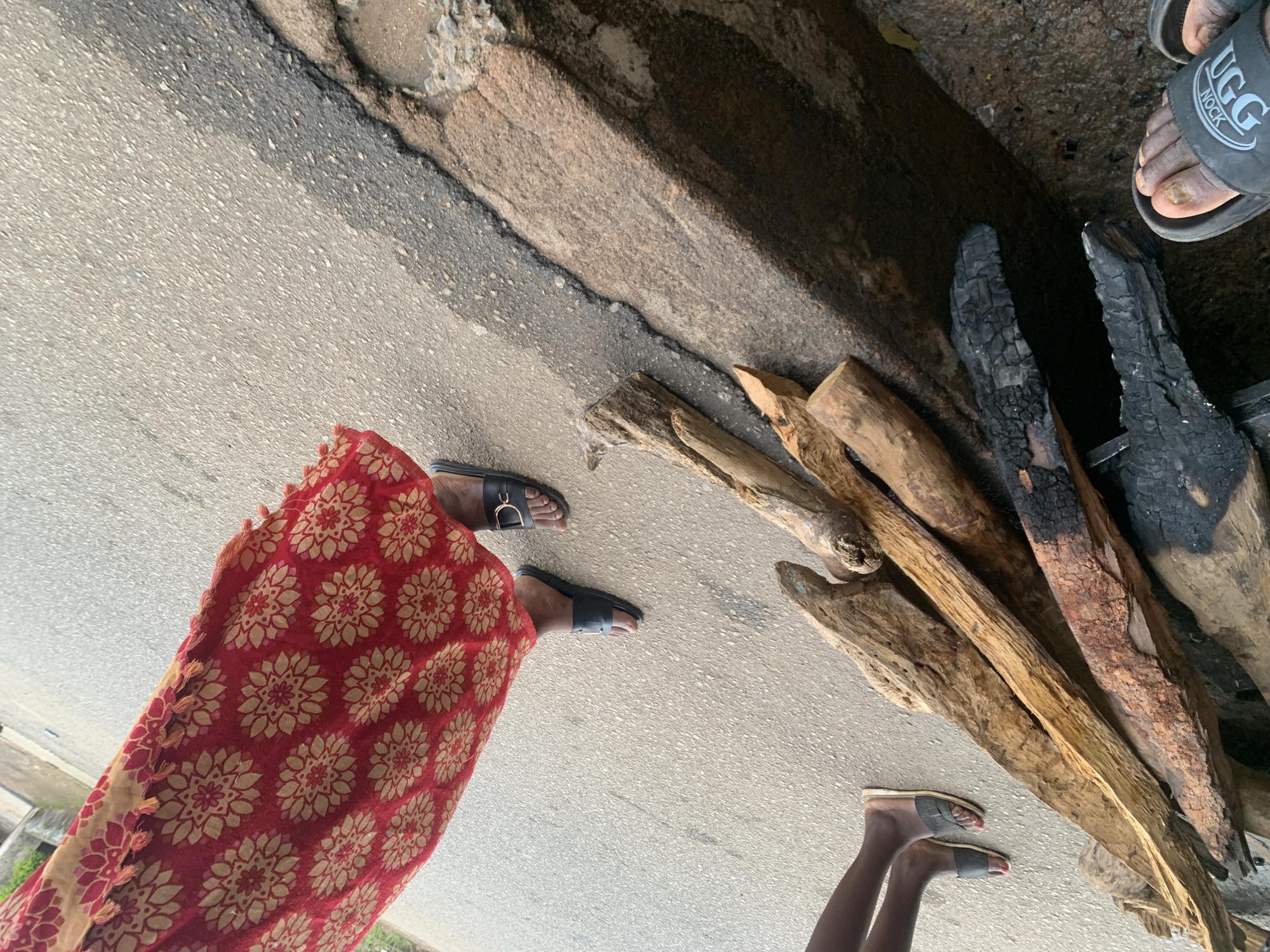 The image depicts a scene outdoors, possibly in Jos, Nigeria, featuring several individuals and objects on a paved surface adjacent to a raised, rough-textured embankment. In the upper left quadrant, a person is visible wearing a red garment adorned with a repeating pattern of floral or star-like motifs in a lighter color, possibly beige or yellow. The hem of this garment is fringed. Next to this person, two feet wearing black slide sandals are visible on the paved ground. One sandal has a decorative metallic element.
To the left of center, another pair of legs, clad in dark skin, is seen wearing grey or dark-colored slide sandals with straps. These feet are positioned on the paved surface.
Dominating the lower half of the image is a pile of logs of varying sizes and conditions. Some logs appear to be light-colored wood, while others are charred and blackened, suggesting they may have been used for fuel or fire. The logs are arranged diagonally across the frame, resting partially on the paved surface and partially on the embankment.
The paved surface appears to be asphalt, showing signs of wear and damage along the edge where it meets the embankment. The embankment itself is rough, rocky, and textured, with some areas appearing wet or stained. The overall lighting suggests it is daytime.