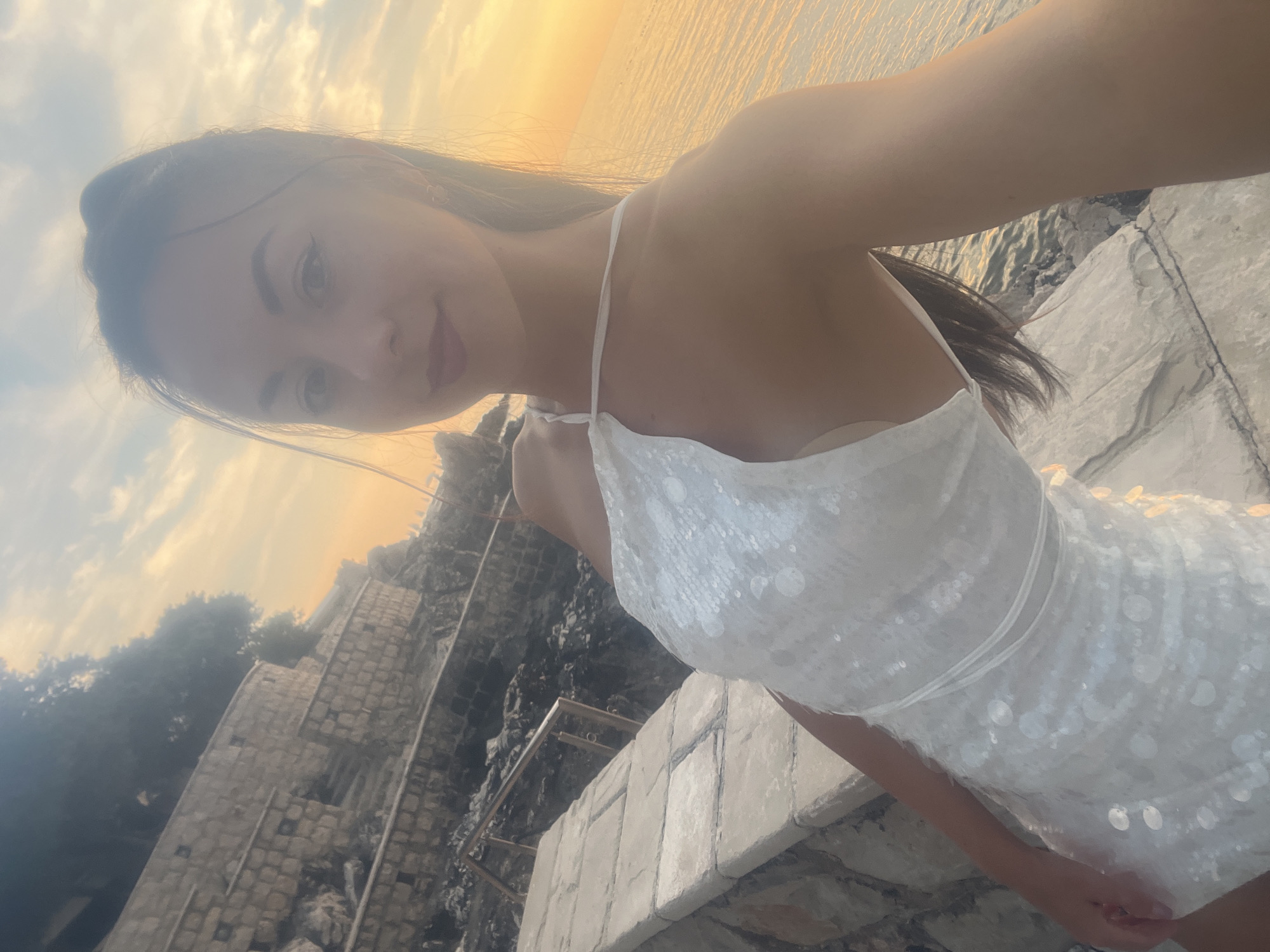 A young woman wearing a white sequined dress is taking a selfie outdoors during sunset. She is standing on a stone ledge by the water, with a rocky incline and trees behind her. The sky is filled with warm, orange and yellow hues from the setting sun, reflecting on the calm sea. The woman has her hair pulled back and is looking towards the camera with a slight smile. The overall scene suggests a peaceful and picturesque coastal setting in Pečurice, Montenegro.