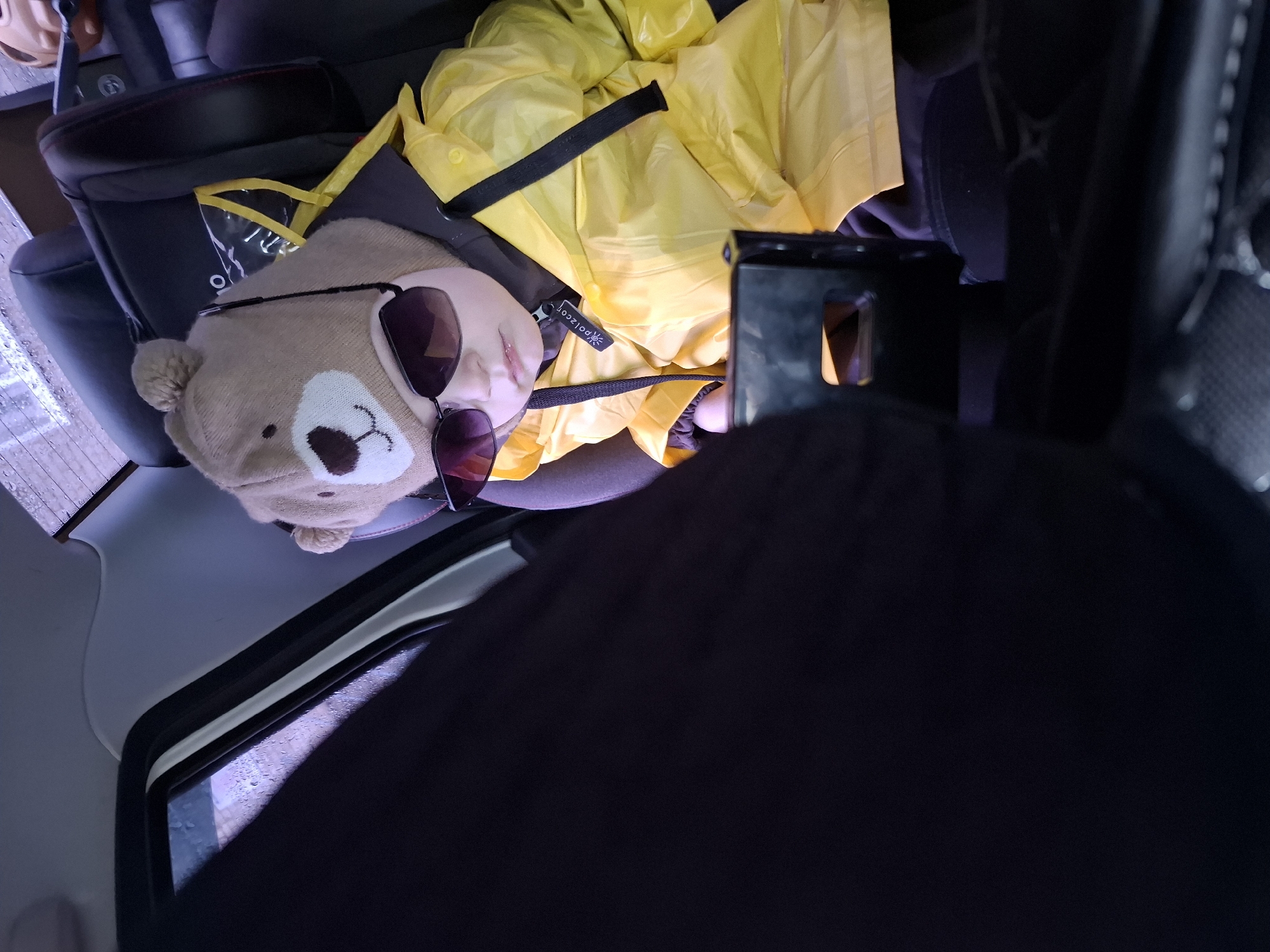 A child in a yellow raincoat and bear-shaped hat with sunglasses rests in a car seat. The scene, likely in a vehicle in Chaikovsky, Russia, shows raindrops on a window. The child is buckled into a dark-colored car seat. The interior of the vehicle is visible with a dark-colored object obstructing a portion of the view.