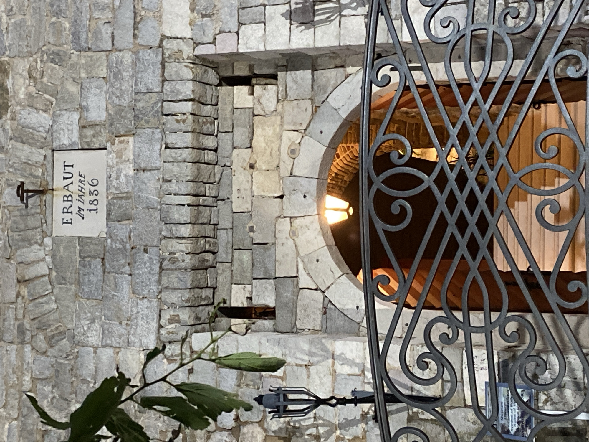 A stone building with a wrought-iron gate in Budva, Montenegro. The building is constructed of light gray stones, irregularly shaped and stacked. A stone plaque on the wall reads "ERBAUT IM JAHRE 1836" in dark lettering. To the right of the plaque, the stone construction changes to stacked flat stones. A circular opening in the wall is partially obstructed by an ornate wrought-iron gate, revealing an interior space with a curved ceiling and a warm light source. A small plant with green leaves is visible in the foreground. A small sign is affixed to the iron gate.