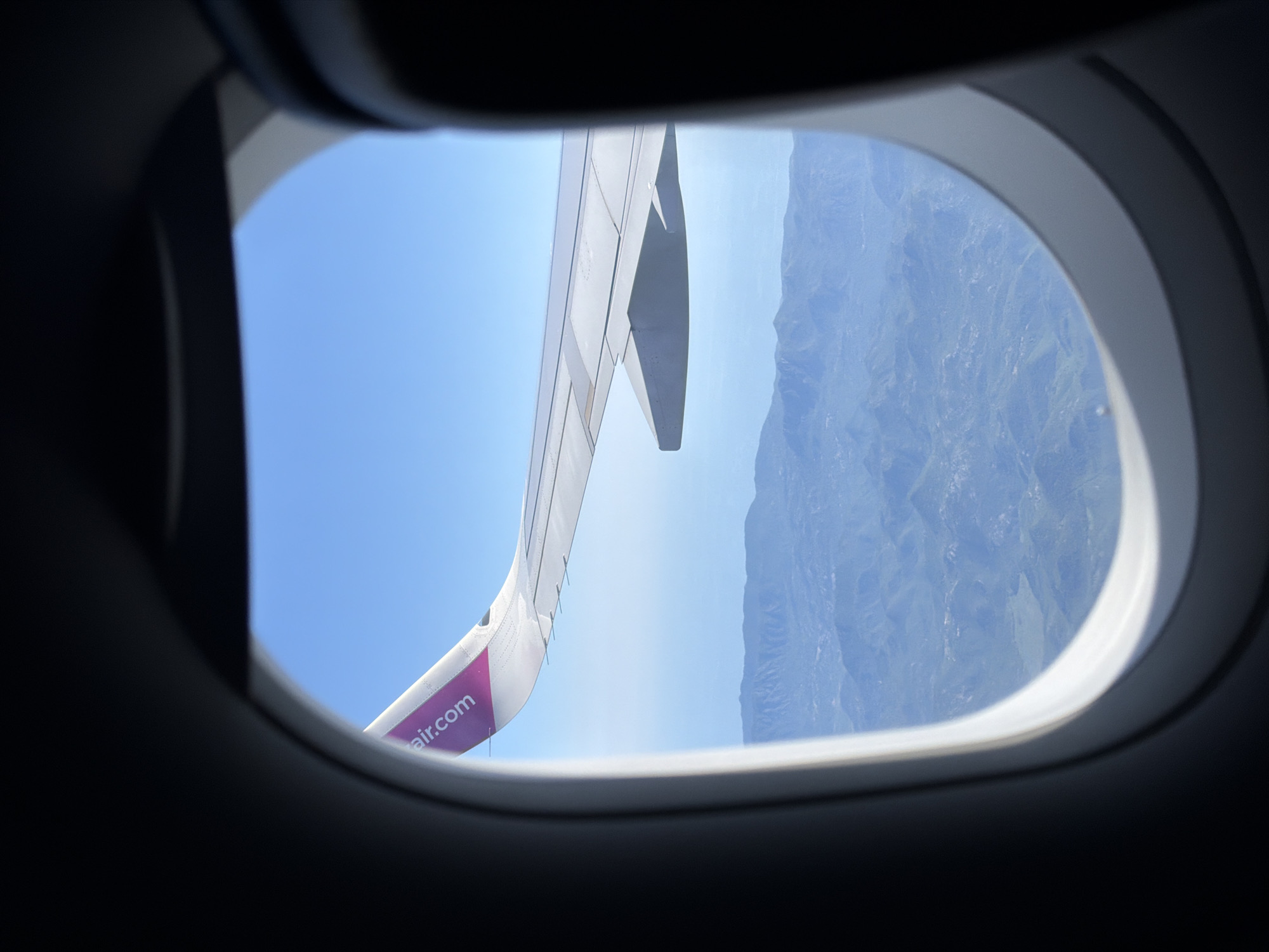 The image shows a view from an airplane window. The window frame is dark, with a bright, oval-shaped opening revealing the sky and landscape below. A portion of an airplane wing, painted white with a magenta logo reading "air.com", extends into the frame from the upper left. The wing is positioned above a vast, mountainous terrain. The mountains appear to be covered in a light green vegetation, with visible valleys and peaks. The sky is clear and a vibrant blue. The scene suggests an aerial view of a rural, possibly coastal, region, consistent with the mountainous landscape of Donja Cijevna, Montenegro.