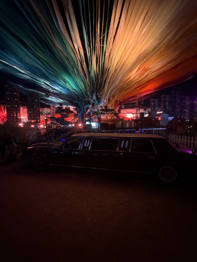 The image shows an outdoor scene, likely at night, in Indio, California, United States. The foreground is a dirt-covered ground leading to a black limousine. The car is parked, and the windows are tinted, making it difficult to see inside. Beyond the car is a structure, perhaps a stage or a building, with an elaborate overhead canopy consisting of draped fabric in a spectrum of colors from blue to orange. Behind this structure, there is a view of what appears to be the city skyline of Indio. The overall lighting is dim, with some artificial lights illuminating the stage area and the city skyline. The environment suggests a temporary or event-related setup.